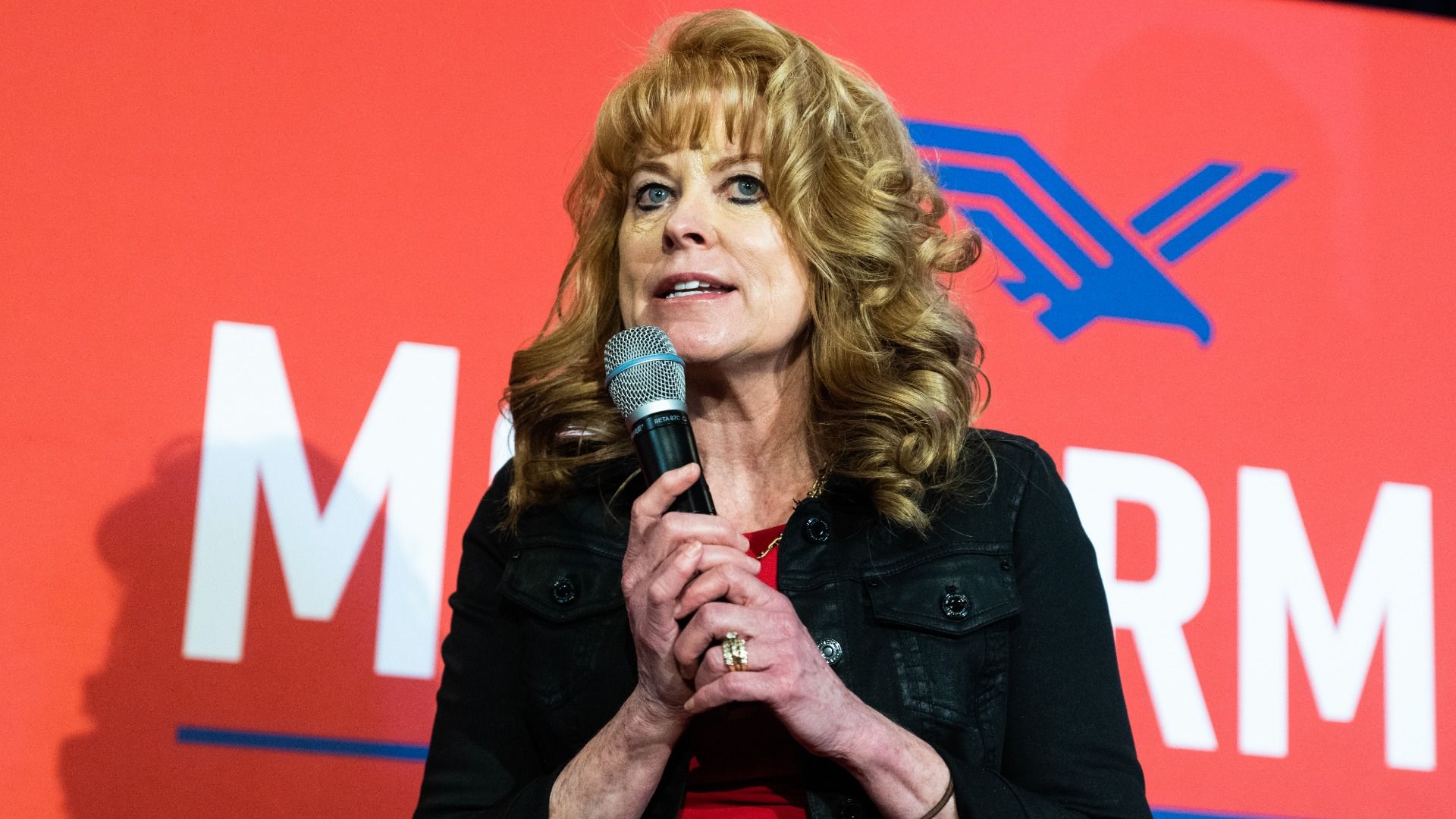 State Treasurer Stacy Garrity holding a microphone while talking in front of a red campaign poster.