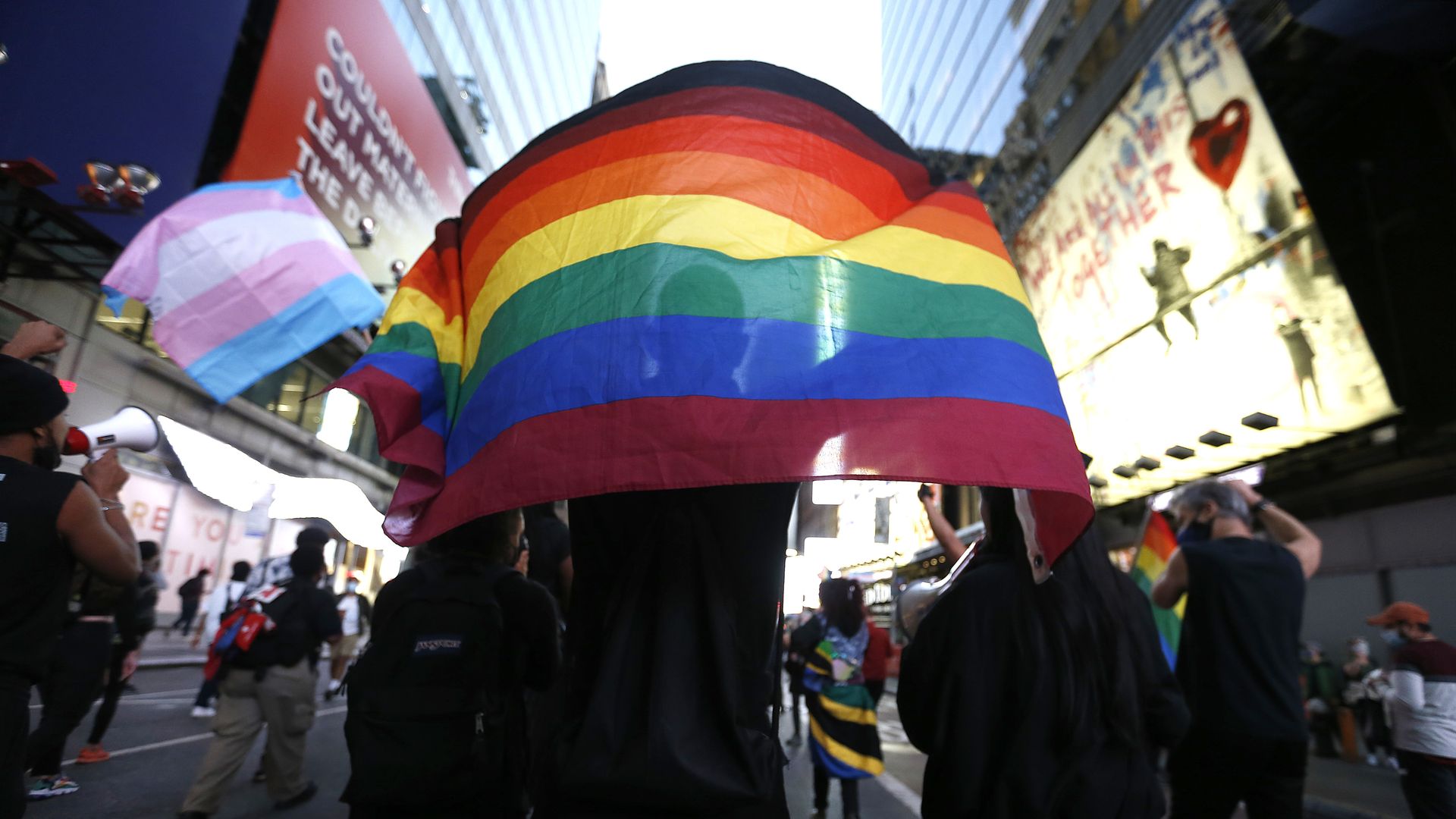 Someone holds a rainbow flag in this image, and someone else holds a trans flag in the background. 