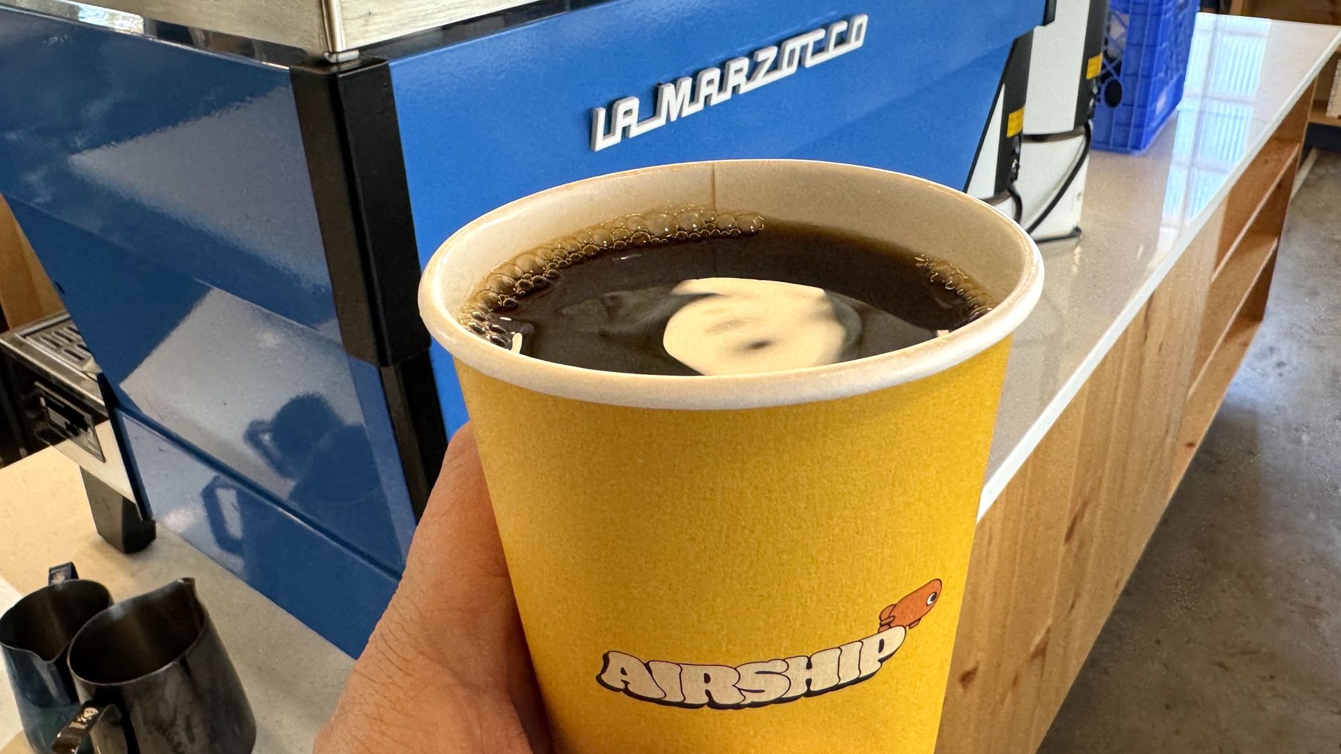 A photo of a cup of coffee in a yellow paper cup held in front of an espresso machine. 