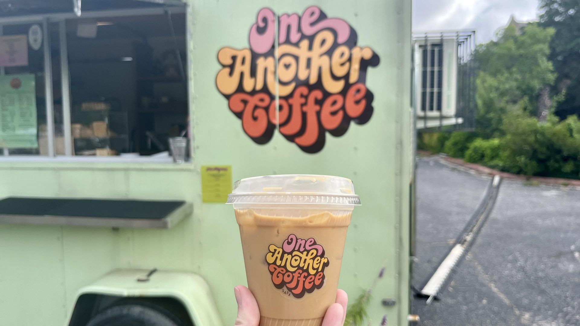 A hand holds an iced coffee in front of a pastel green food truck. Both the cup and truck feature the colorful "One Another Coffee" logo in retro bubble letters. The truck window is open, with greenery and a cloudy sky in the background.