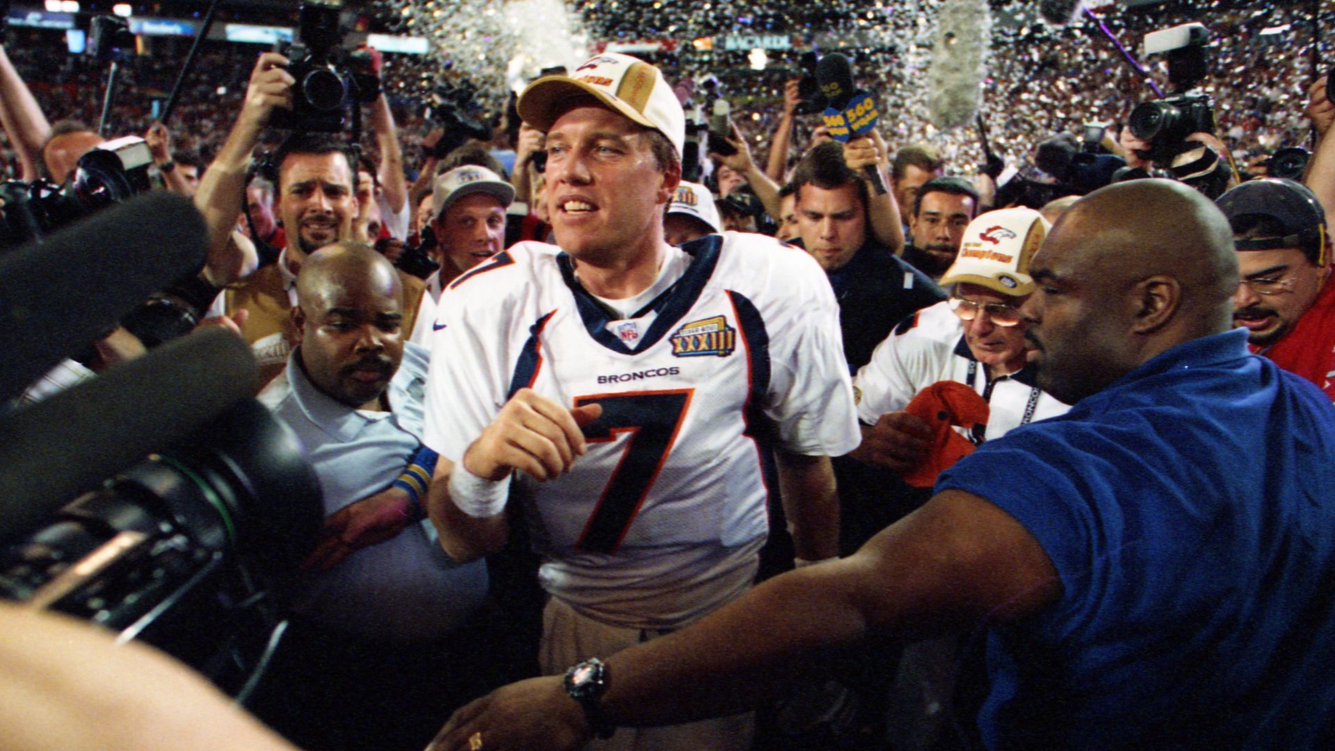 Denver Broncos player wearing number 7 jersey celebrating on the field surrounded by crowd and media with cameras and microphones, confetti falling in the background.