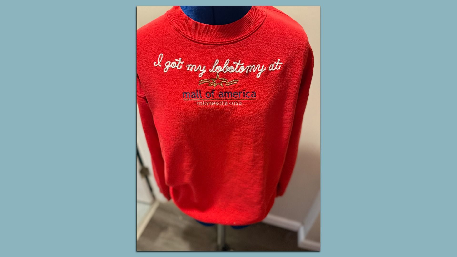 Red sweatshirt with white and navy text reading, "I got my lobotomy at mall of america minnesota usa," displayed on a mannequin indoors.