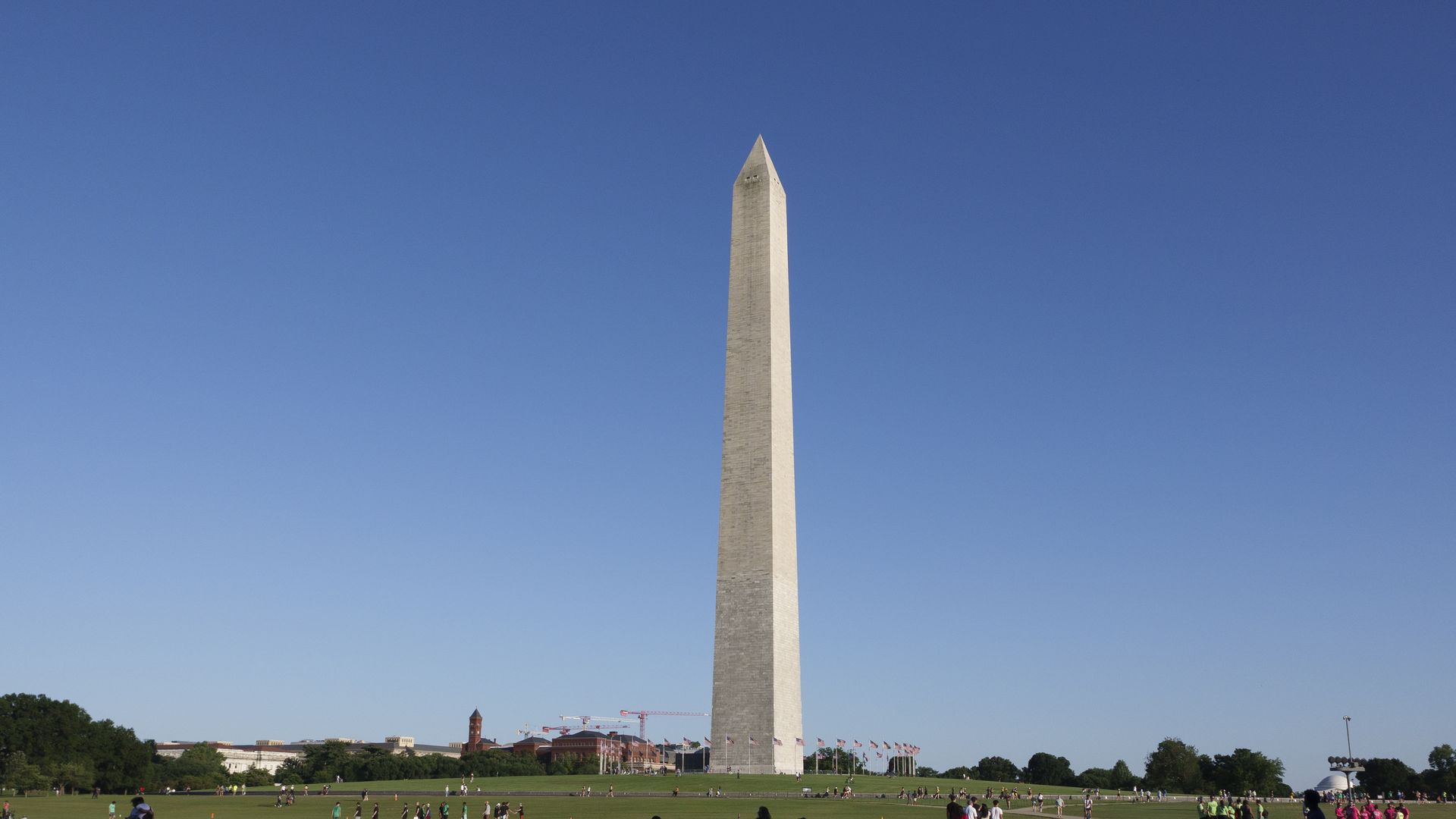 Washington Monument obelisk rises against a clear blue sky; a grassy plaza with visitors, a rope barrier at the base, and flags in the background with distant buildings visible.