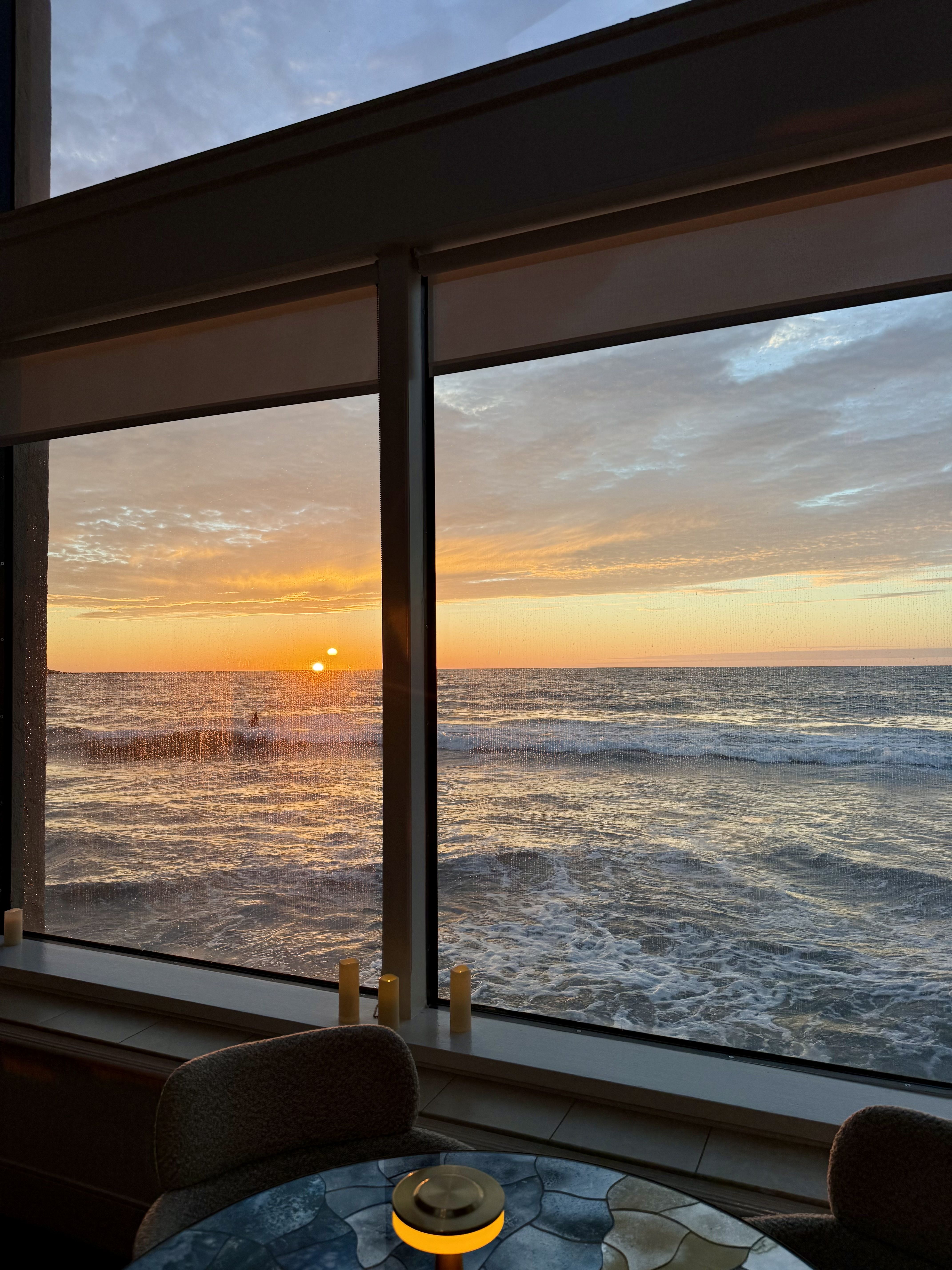 Interior with a large window looking out to a sunset over the ocean. Warm light from candles on the windowsill, while a glass mosaic table and two chairs sit by the view.