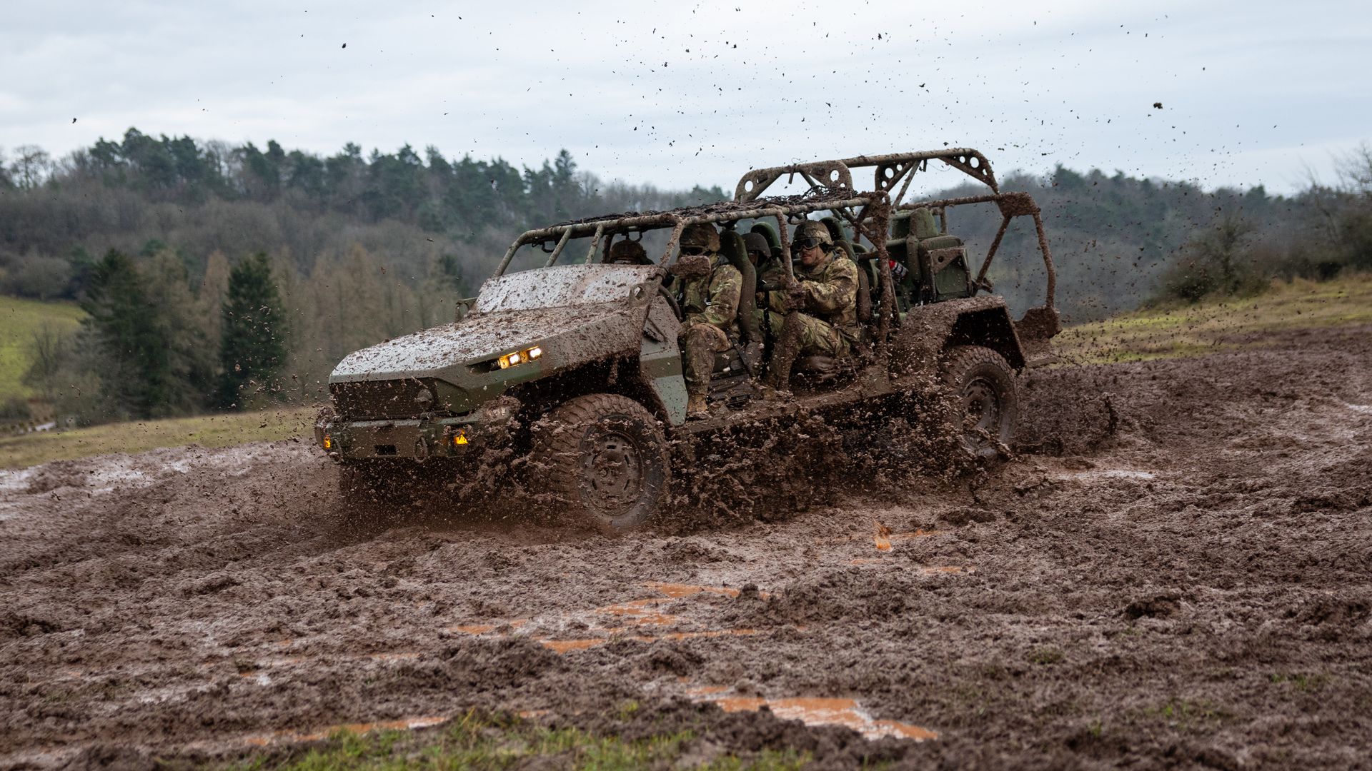 A military vehicle carrying at least three U.S. soldiers tears through a mud pit in forested area.
