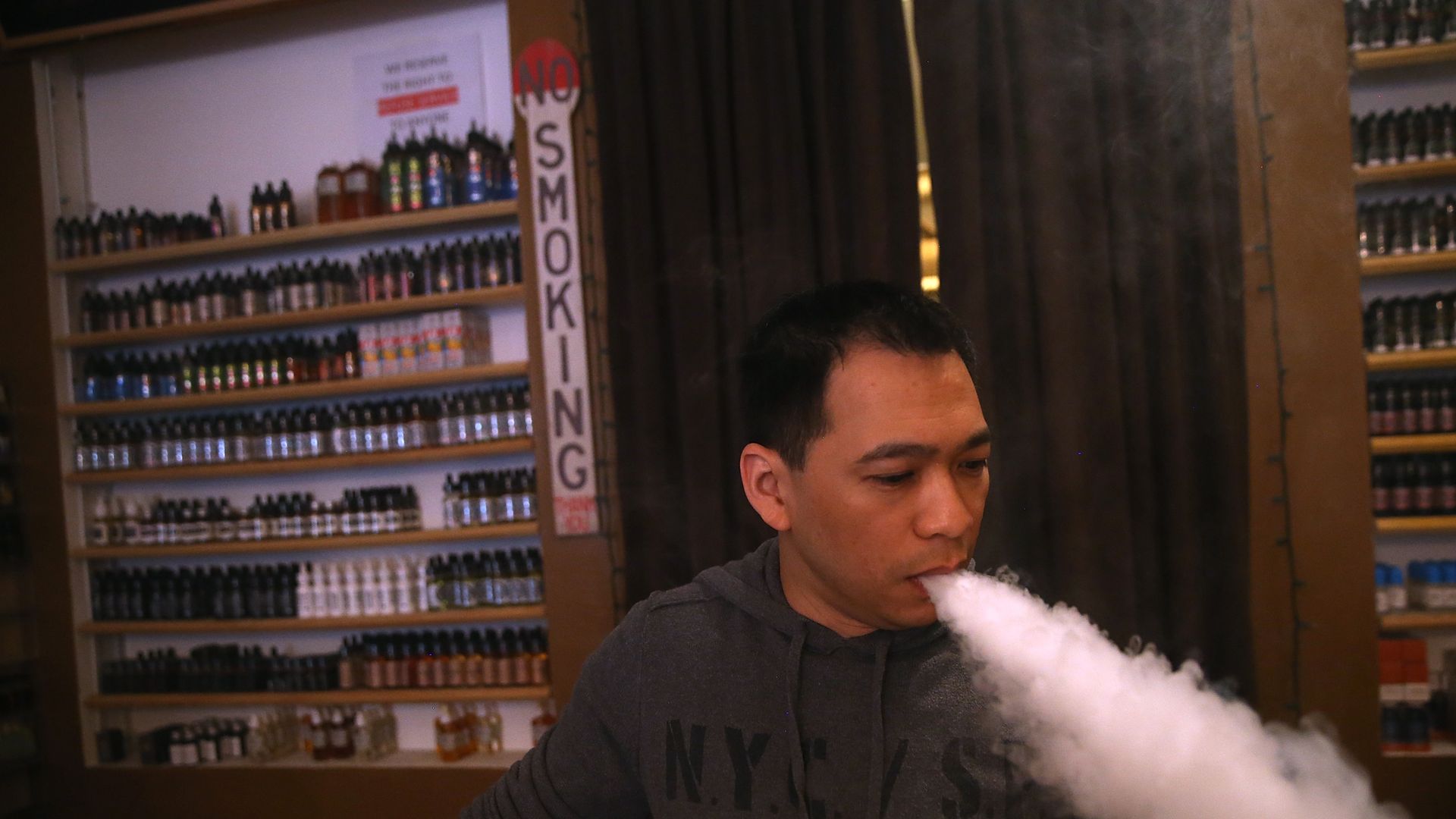 In this image, a man blows a cloud of white smoke down and behind him a shelf of vaping products says "no smoking."