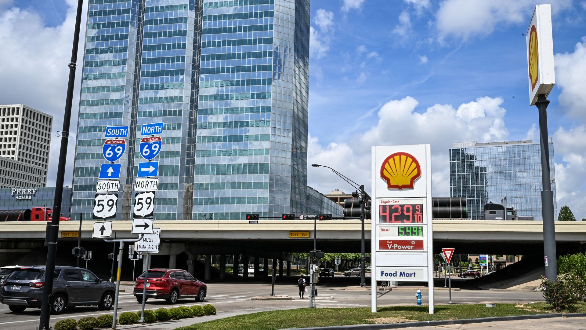 A city street with a tall glass office tower, an overpass, and a Shell sign in the foreground. Blue highway signs show I-69 South/North and US 59; cars and a pedestrian nearby.