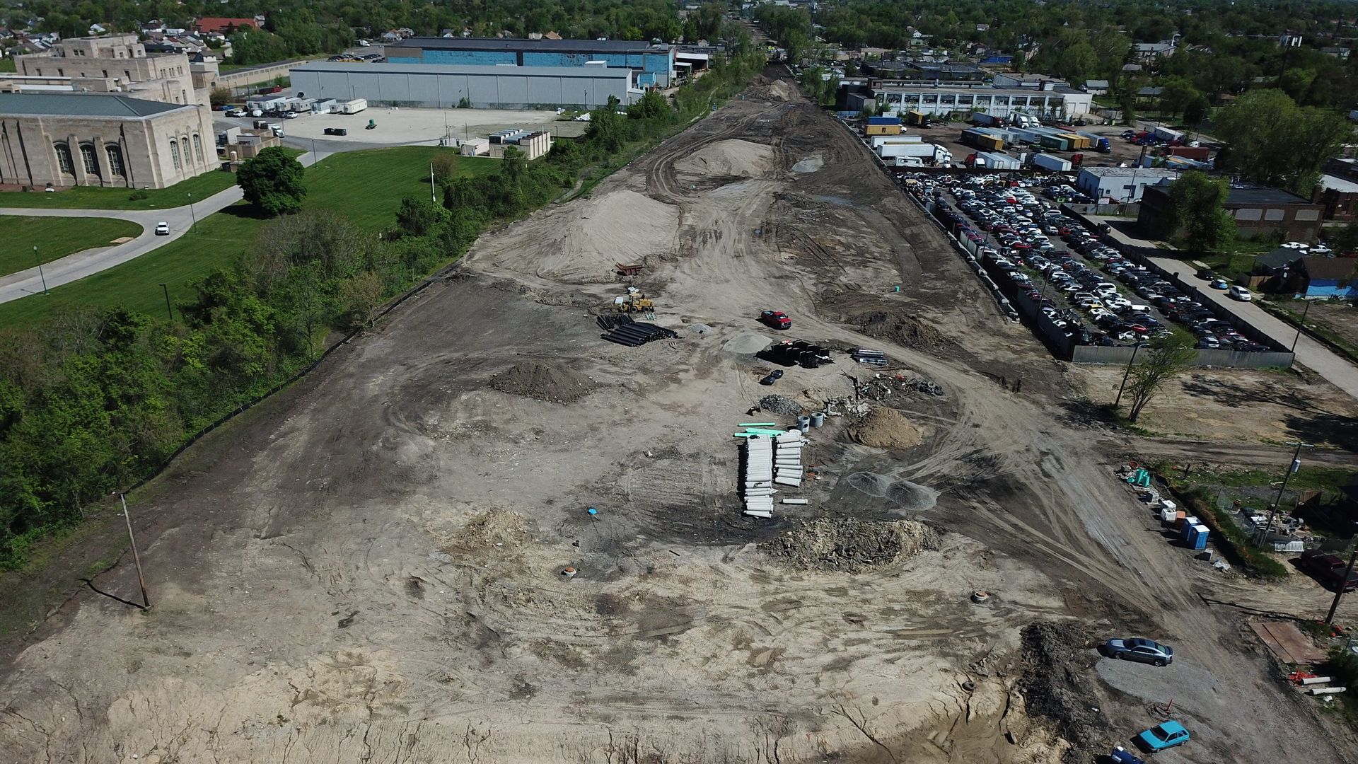 A long construction area is shown from above, surrounded by buildings and trees.