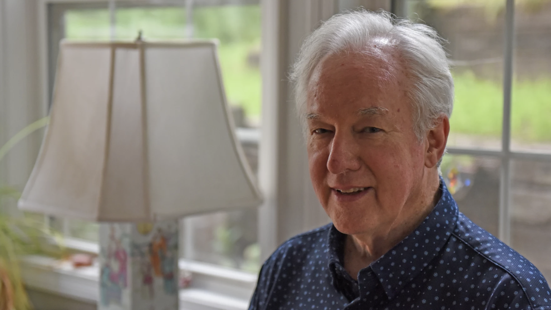 An elderly man with white hair sits indoors beside a decorative lamp, wearing a dark blue polka-dotted shirt; a window behind shows green outdoor scenery.
