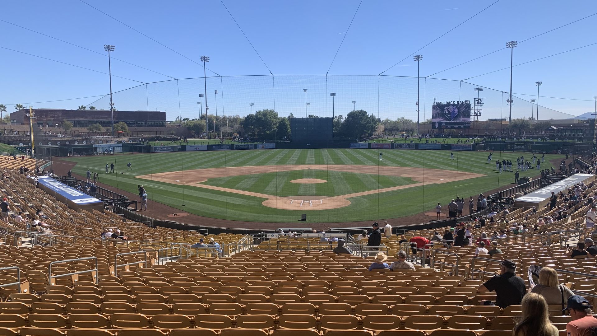 Wide view of a sunny baseball stadium with empty yellow seats in the foreground and players warming up on the green field under a clear blue sky.