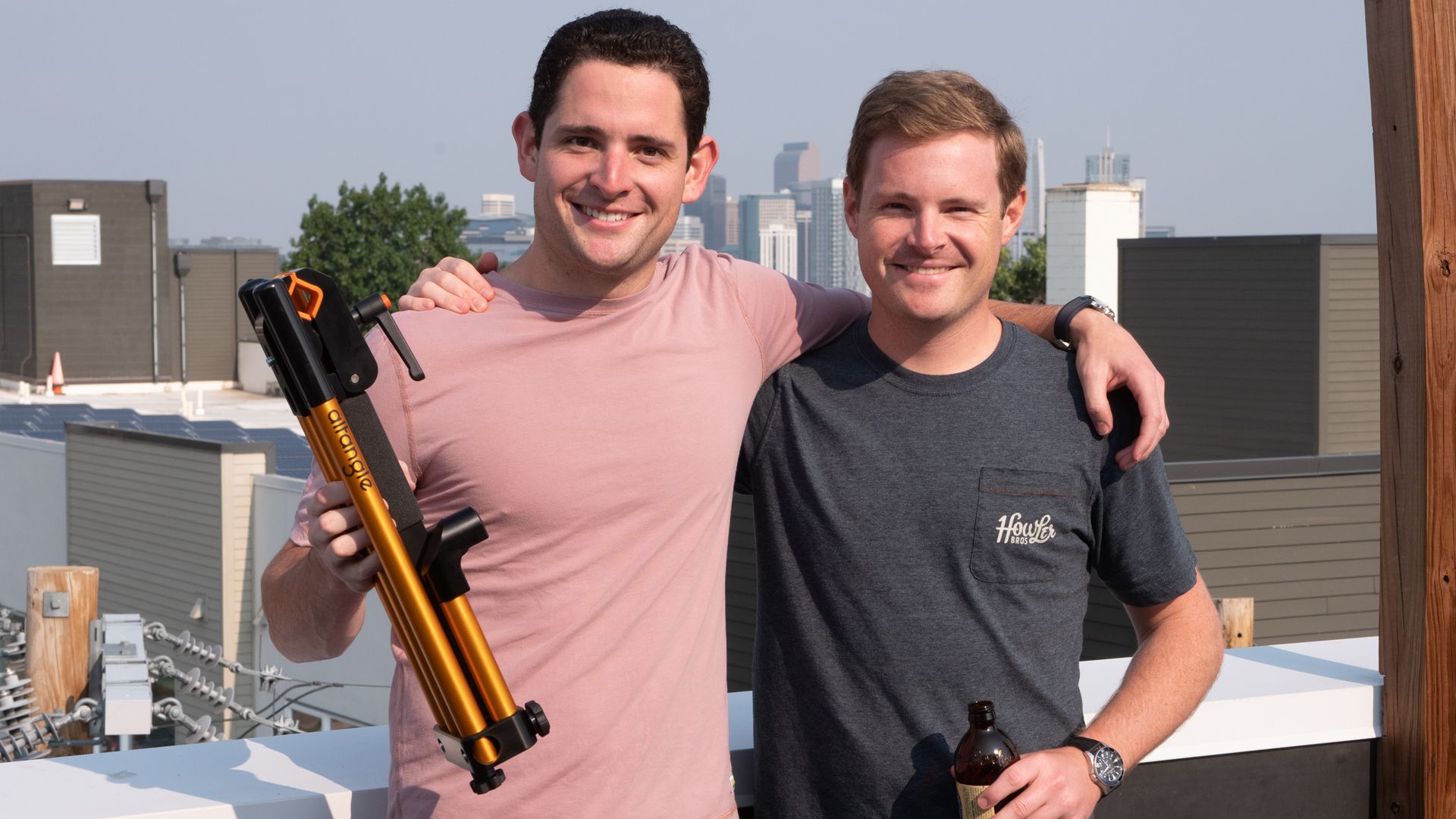 Two men standing side-by-side, one with his arm around the other; one holding an orange tubular contraption and the other a bottle of beer. 