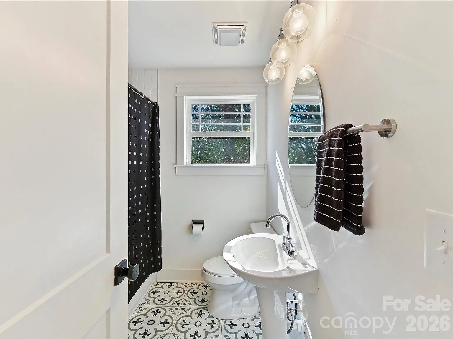 Small bathroom with white walls, patterned black-and-white tile floor, white pedestal sink, oval mirror, black towel with white stripes, black shower curtain with white dots, and window showing trees.