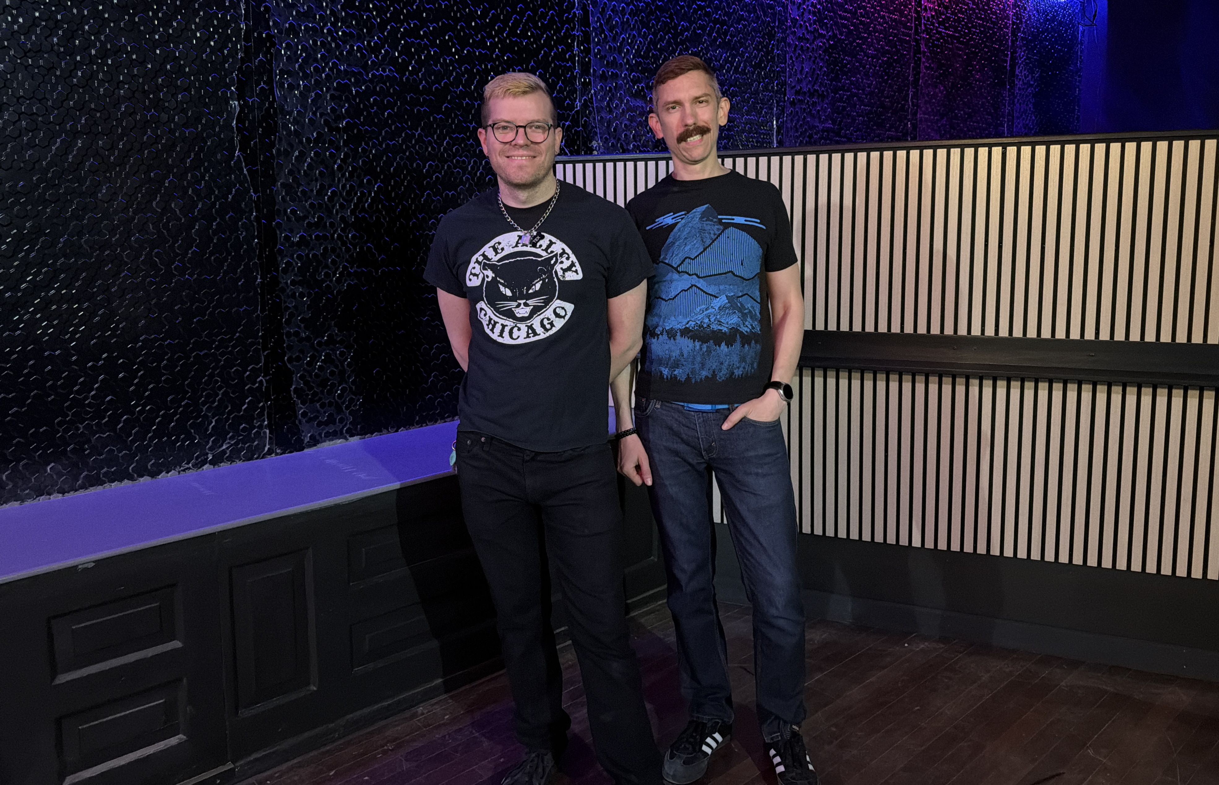 Two men stand together in a dim venue with purple lighting. Left: glasses, blond hair, black T‑shirt with a circular cat logo. Right: mustache, blue mountain graphic T‑shirt; both wear jeans.