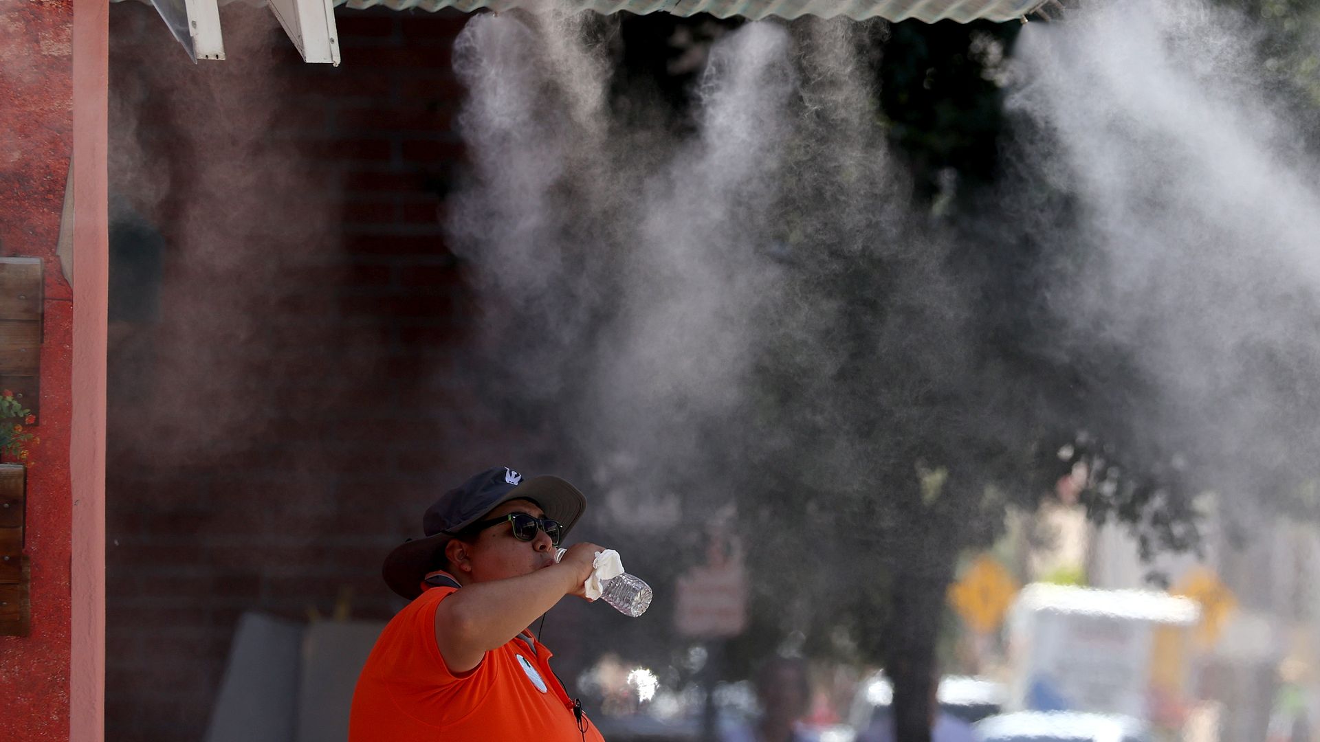 Person in an orange shirt and hat, wearing sunglasses, drinks from a water bottle beside an orange sign reading "Handmade Vodka" as mist swirls in the air.