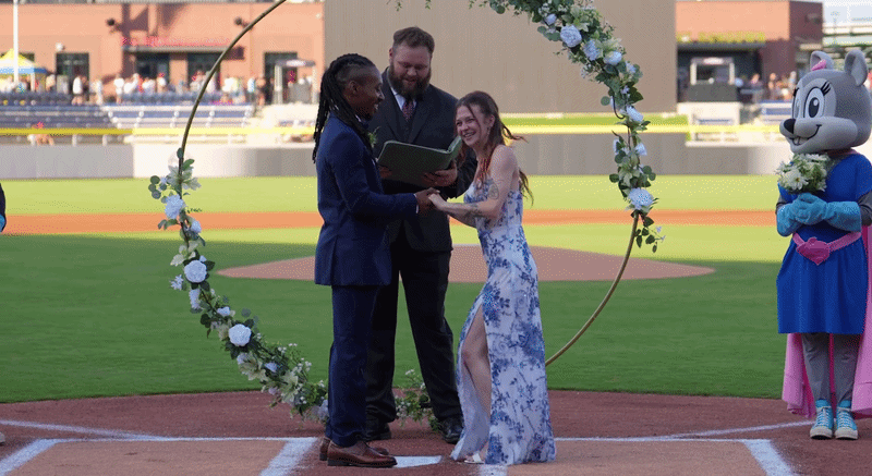 a video of a couple getting married on a baseball field. the bridge jumps up and down before the groom kisses her. A flying squirrel mascot is off to the side with a bouquet