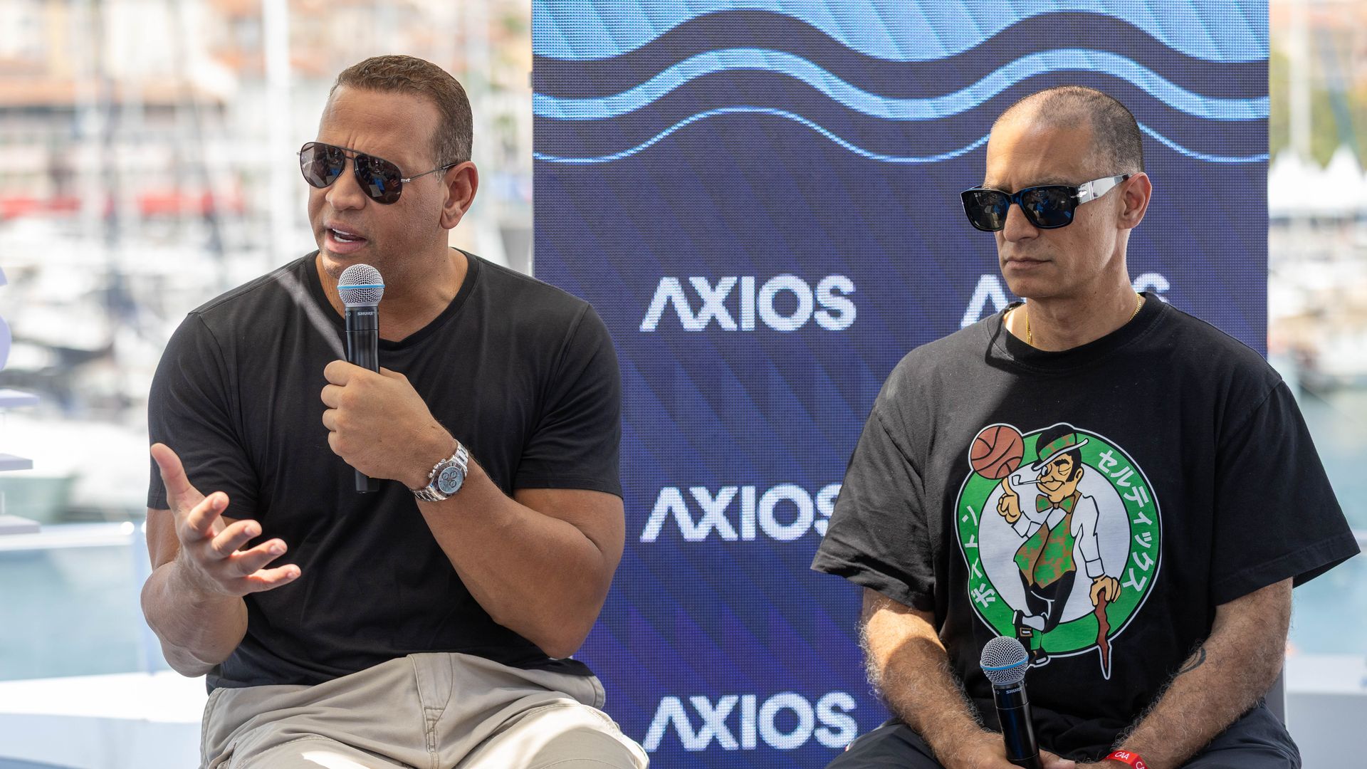Two men in sunglasses speak into handheld microphones on a stage, with a blue Axios banner featuring waves in the background