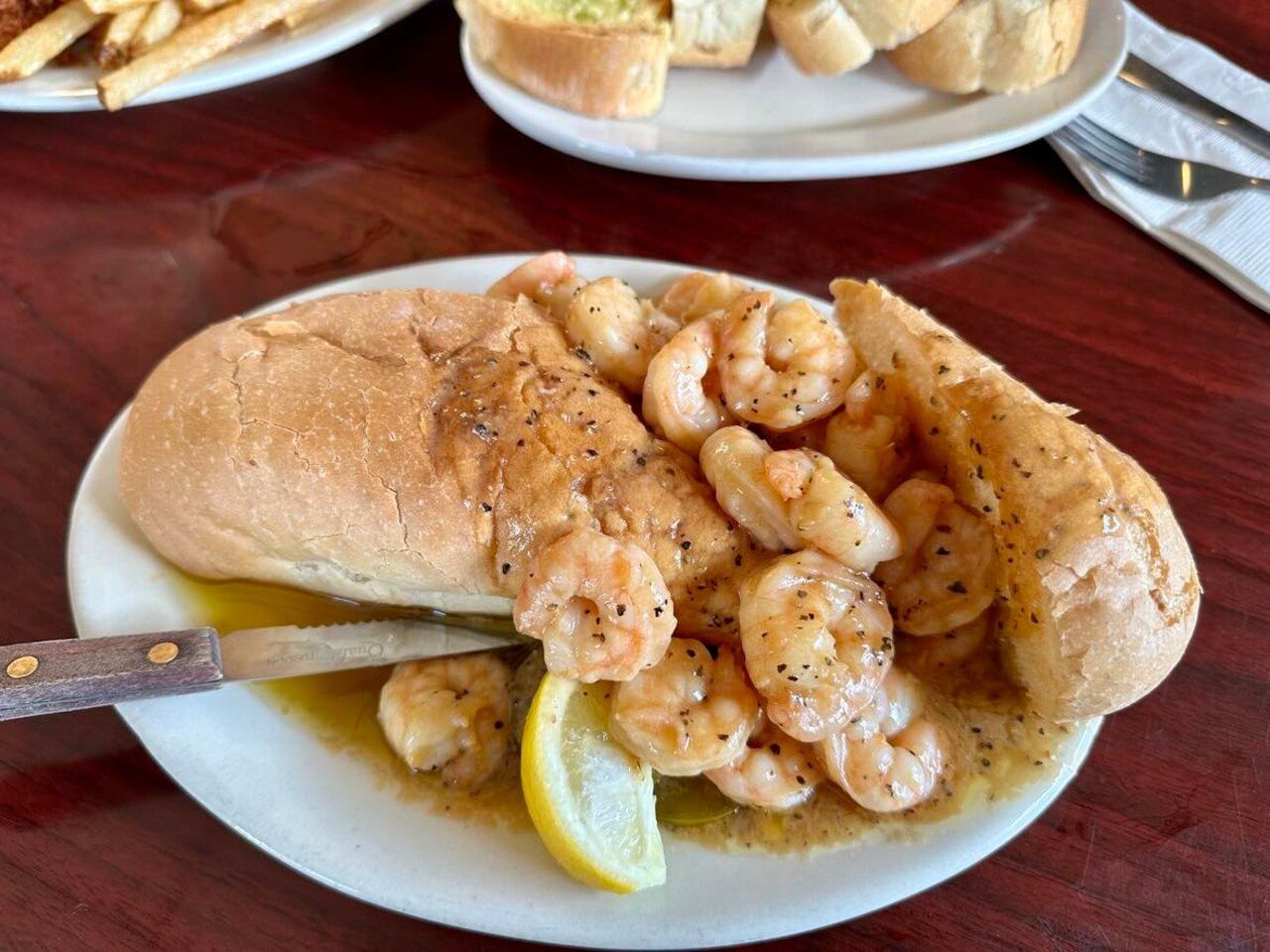 Photo shows a BBQ shrimp poboy on a white plate with a lemon wedge and buttery sauce covering everything.