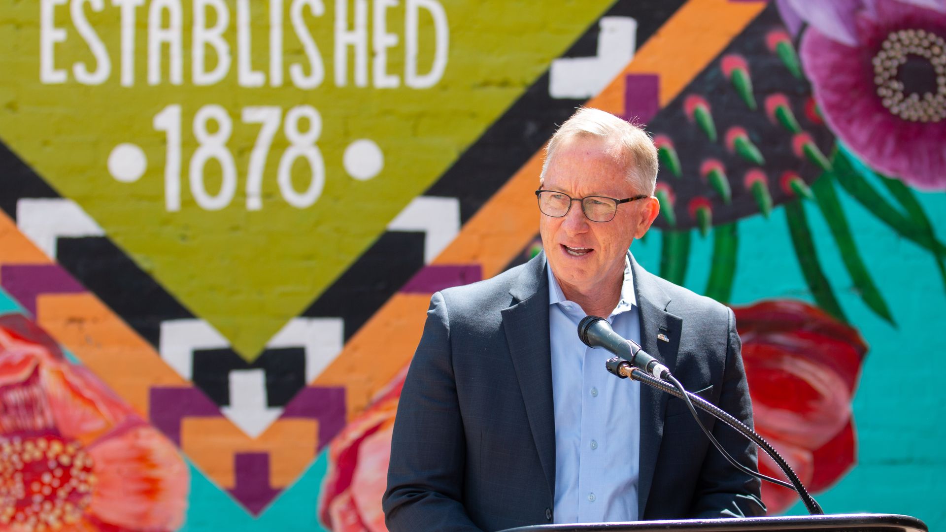 A man speaking at a podium in front of a brightly colored mural.