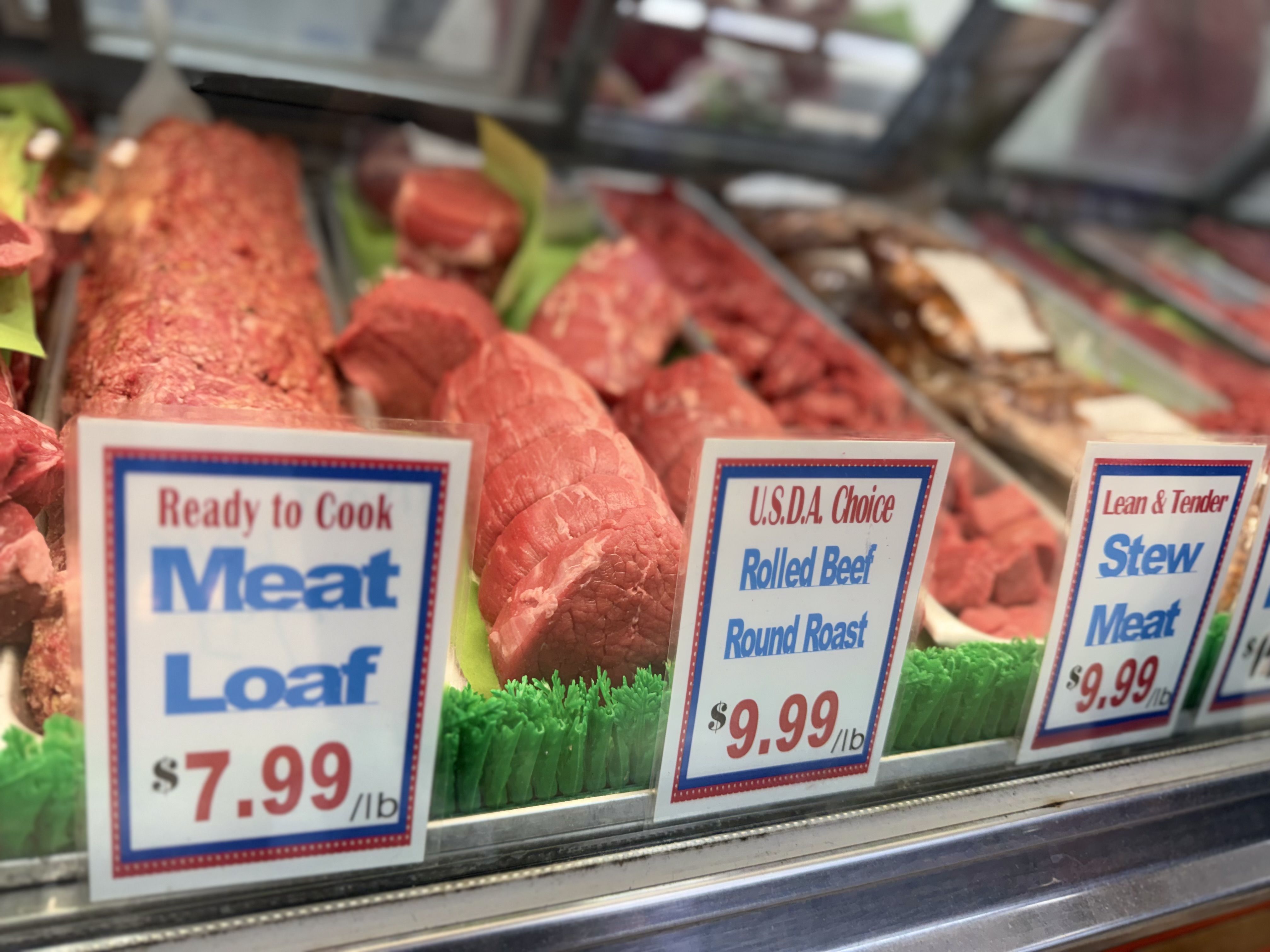 Blurry view of a meat counter: raw beef in trays behind glass, red cuts with blue and red signs reading USDA Choice and Lean & Tender, with prices visible.