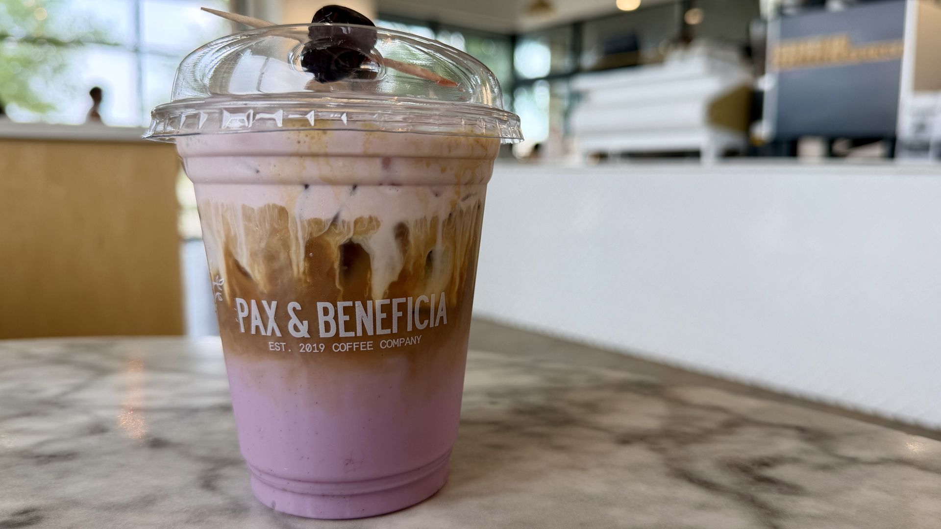 Close-up of a pink and brown layered iced drink in a clear plastic cup with a domed lid, a wooden stirrer and an olive garnish; the cup shows "PAX & BENEFICIA".