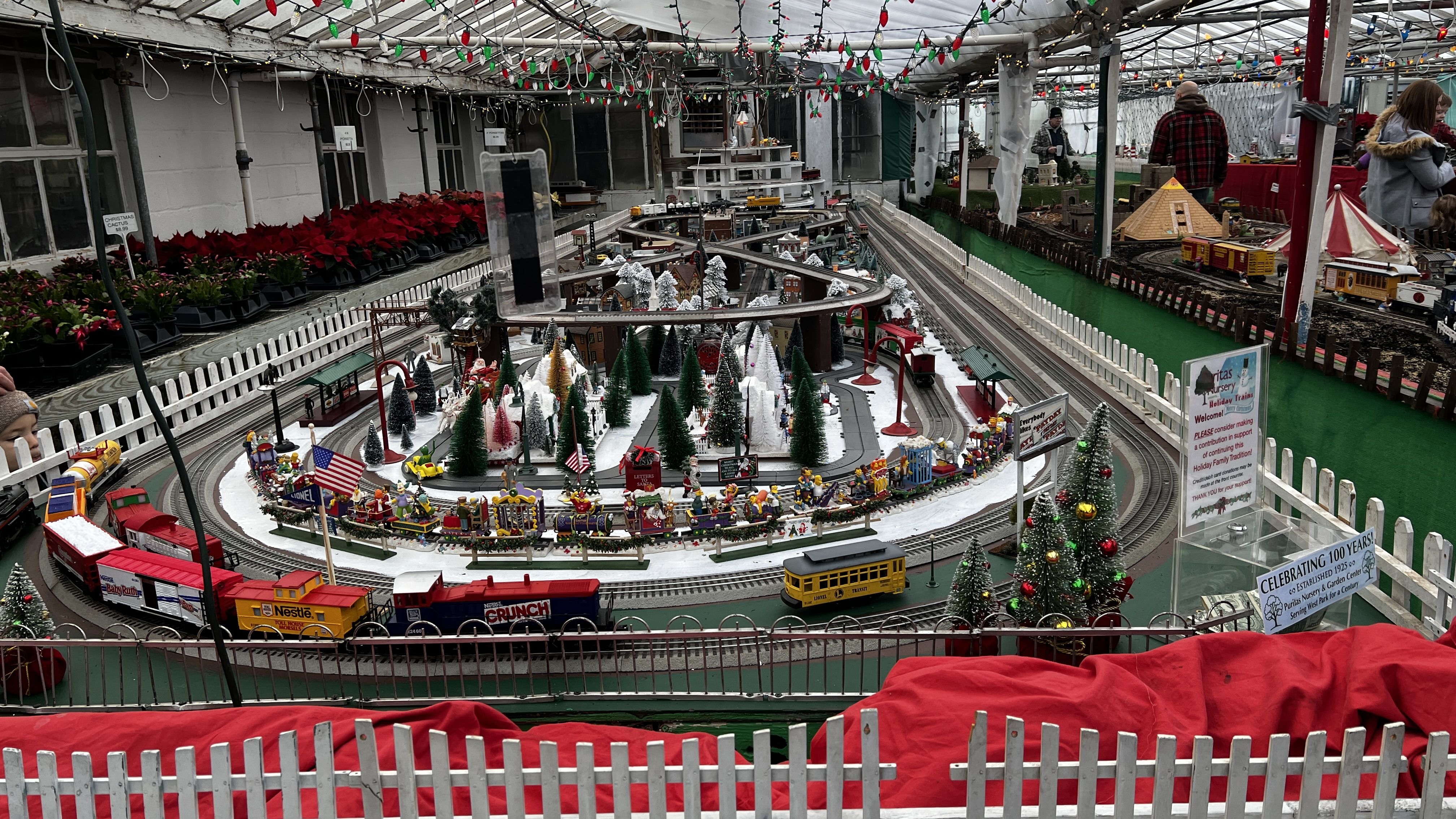 Indoor holiday train display with colorful toy trains, miniature Christmas trees, and snowy miniature village surrounded by white picket fence and festive string lights.