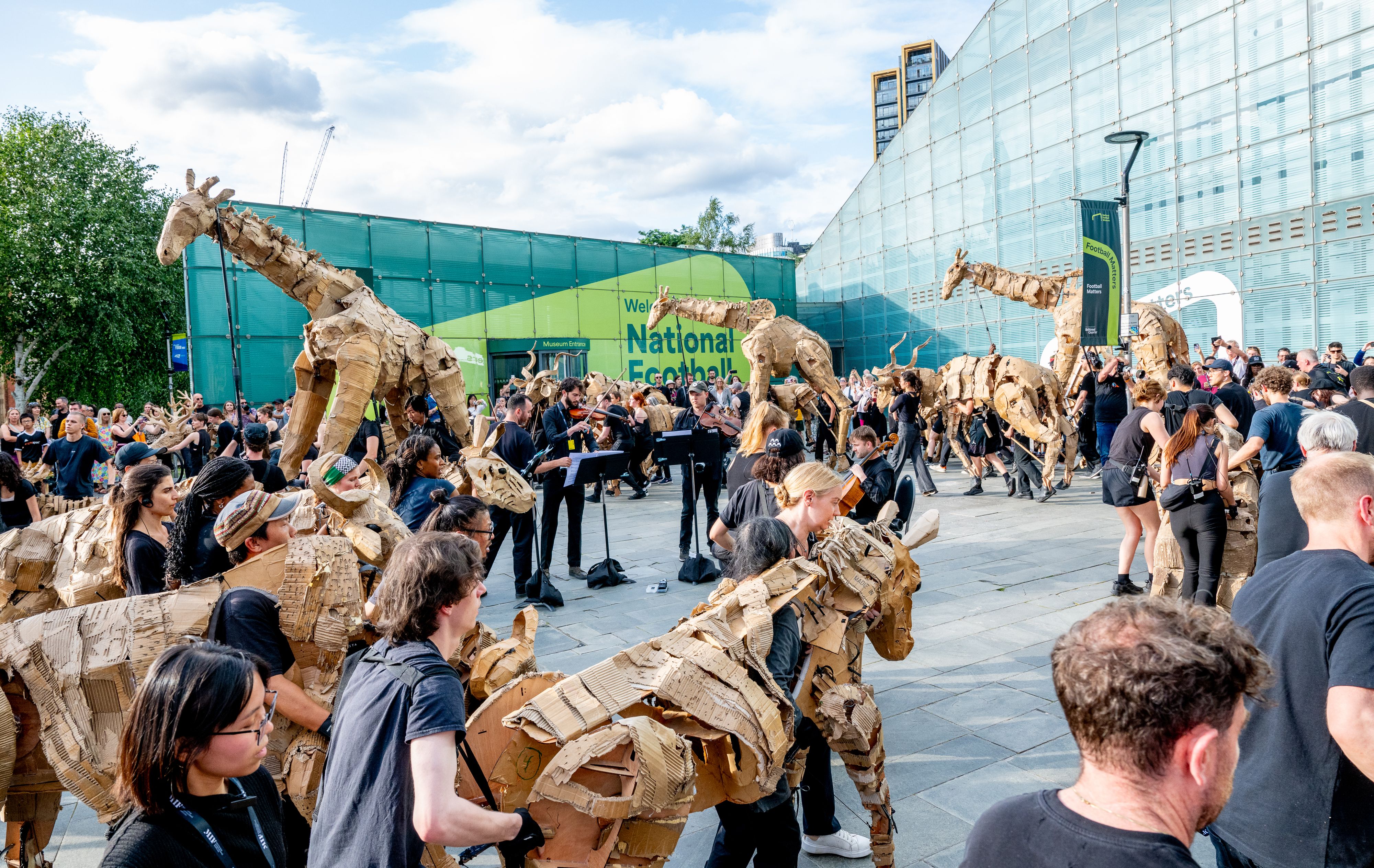 Crowd of people outdoors operating large animal puppets made of cardboard, including giraffes and antelopes, with musicians playing string instruments nearby under a partly cloudy sky.
