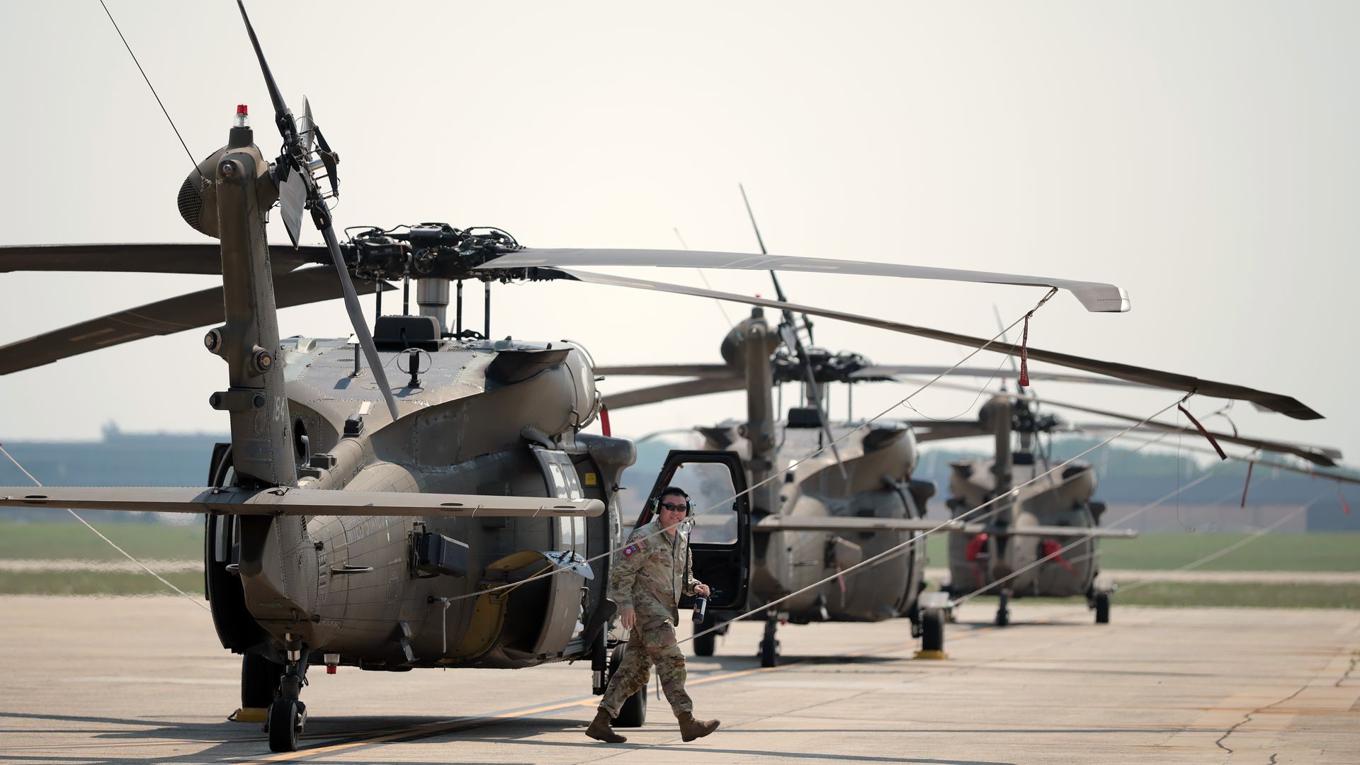 A row of gray military helicopters with large rotor blades parked on a sunlit airstrip; a soldier in desert camouflage walks away from the nearest helicopter, its door open.