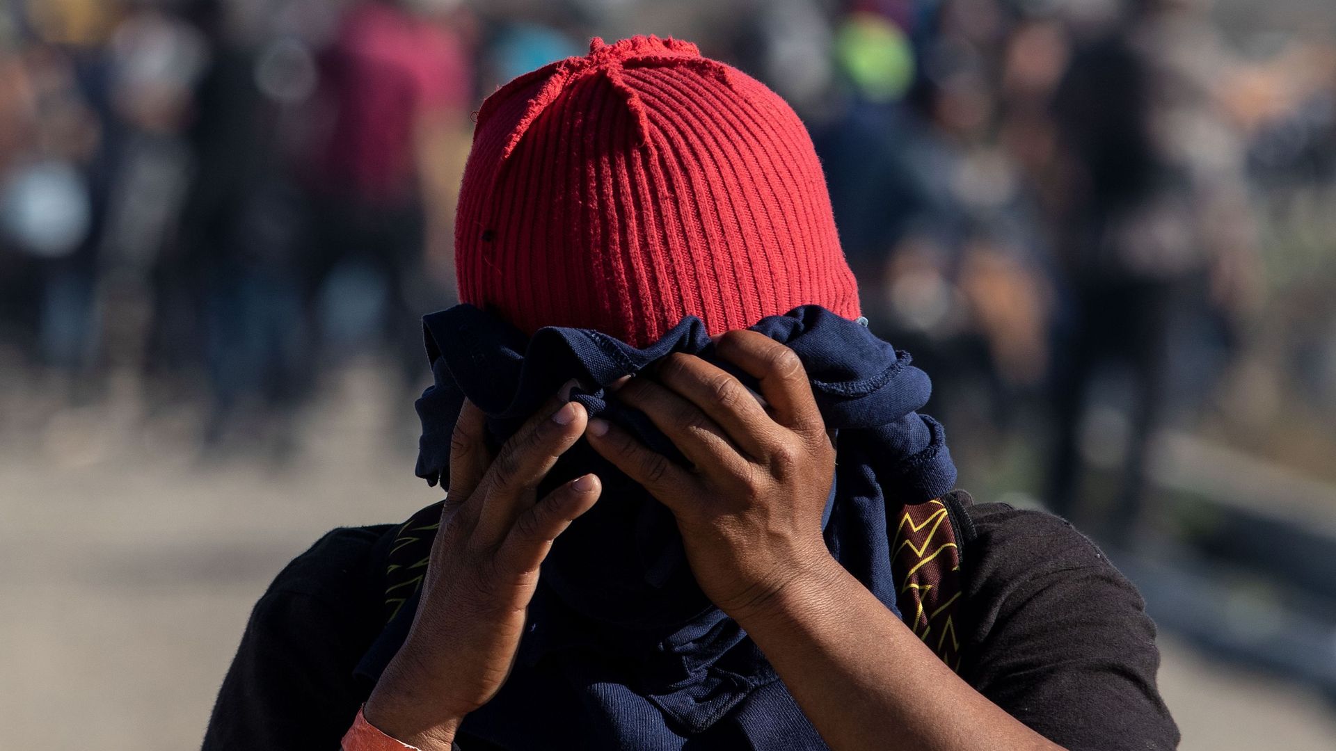 A close up shot of a migrant wearing a red hat and using a navy blue t-shirt to cover their face as tear gas is launched from U.S. border agents. 