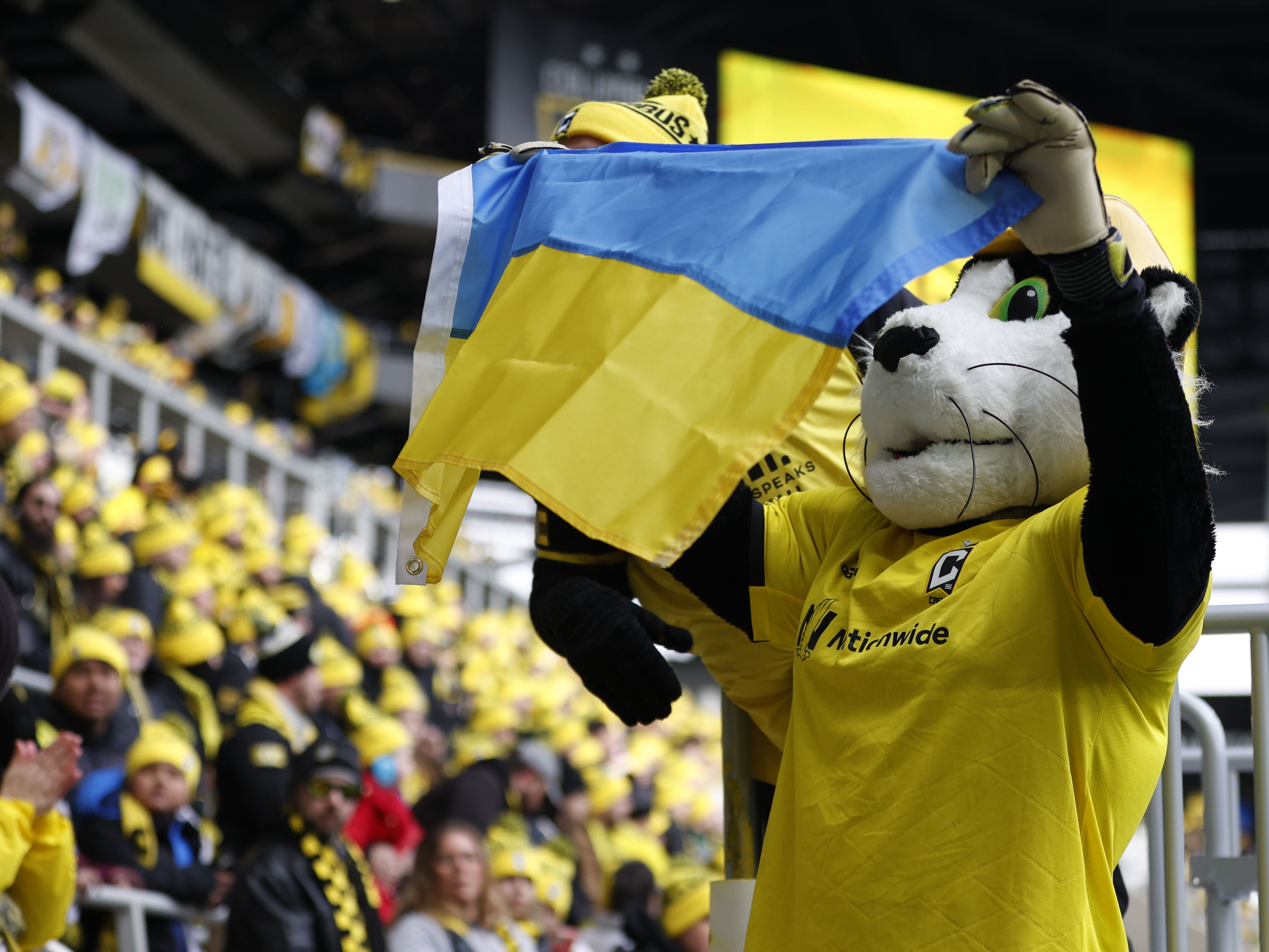Sports mascot in yellow jersey holds a blue and yellow Ukrainian flag at a stadium filled with fans wearing yellow hats and scarves.