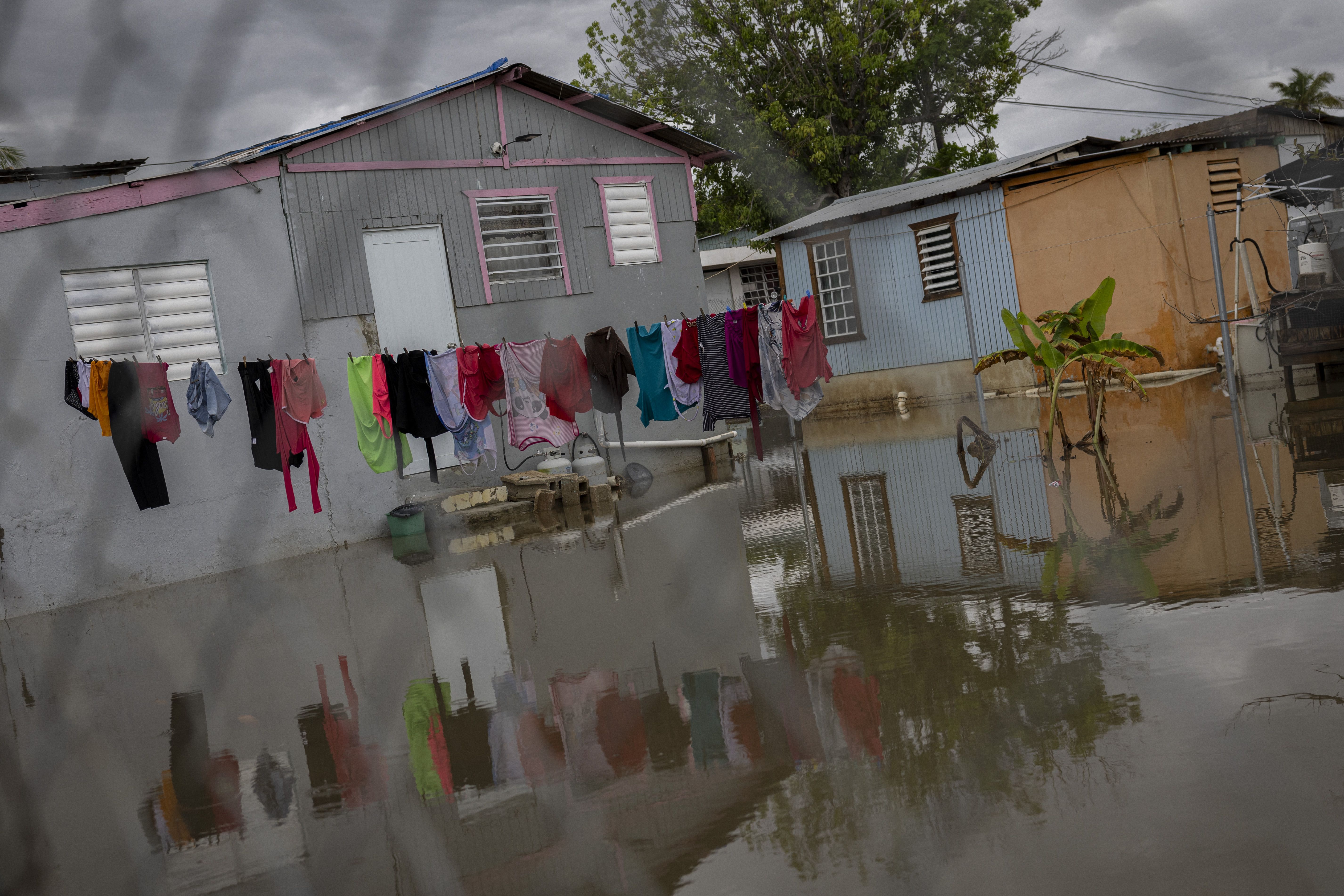 Flooded area with murky water reaching the walls of gray and orange houses; colorful clothes hang on a line above the water under a cloudy sky.