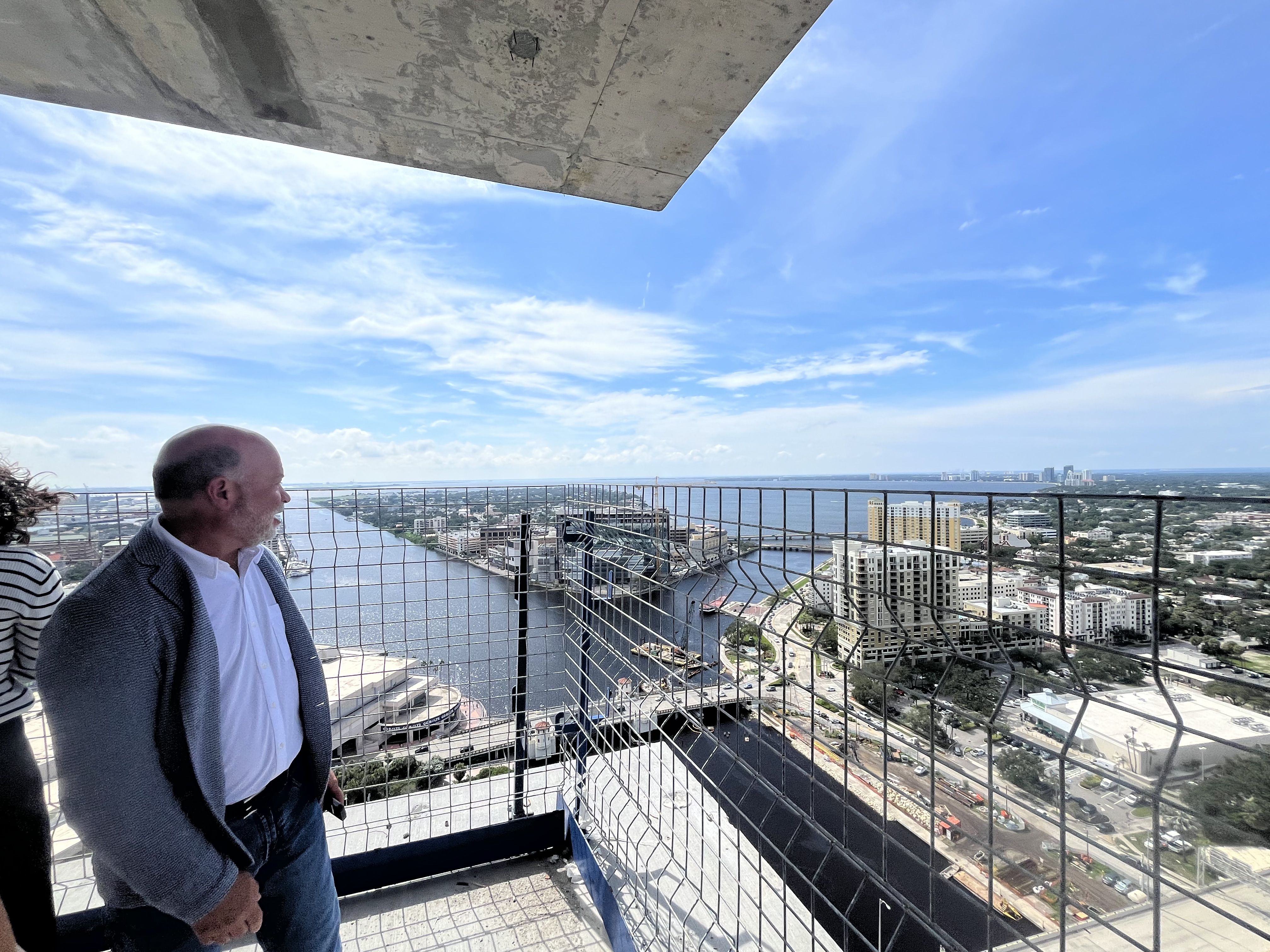 Man in a blazer and white shirt stands on a rooftop balcony with a metal mesh railing, gazing toward a river and city skyline beneath a blue sky with wispy clouds; a concrete overhang looms above.