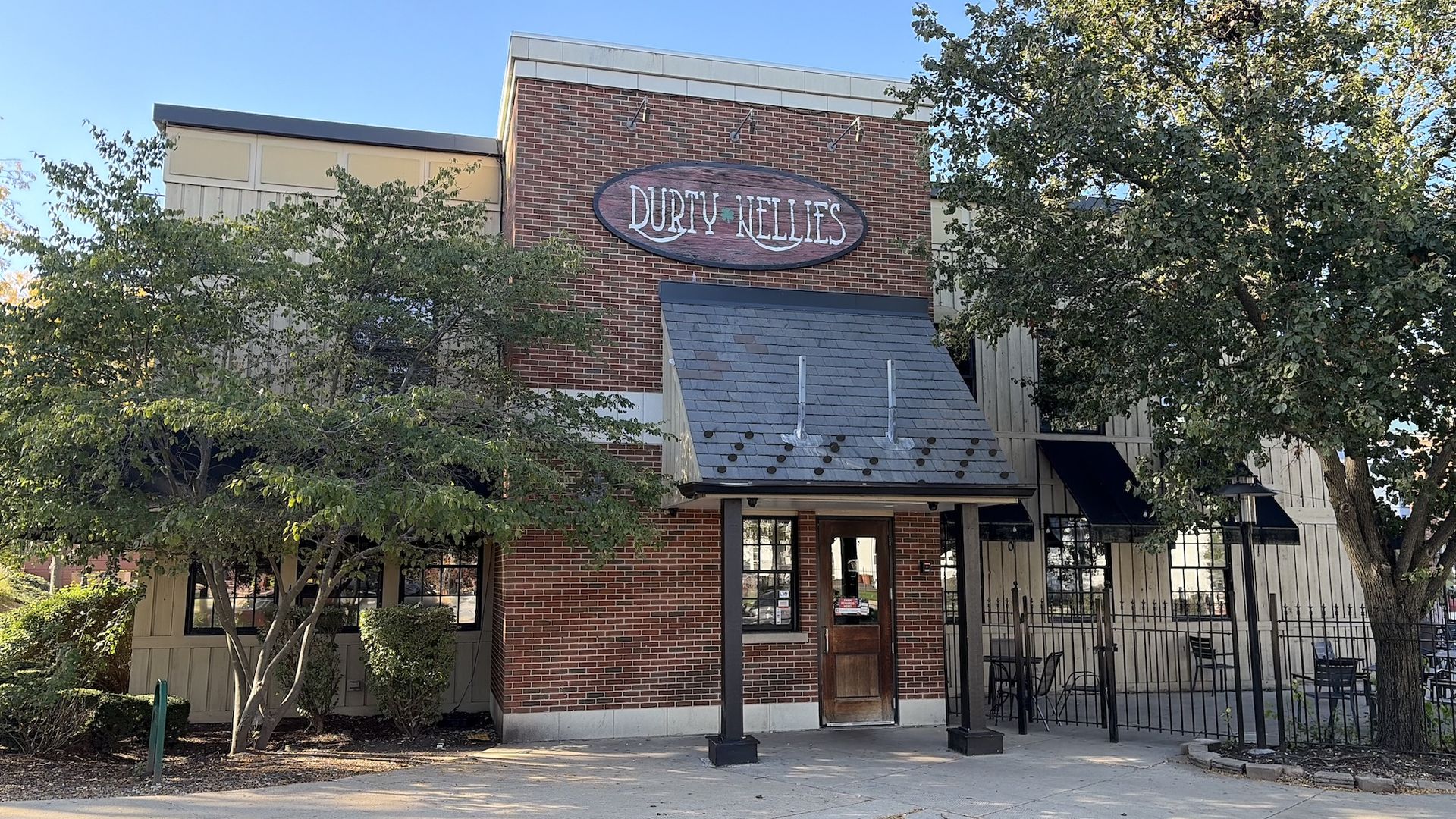 Sunny street scene of a brick building with an oval sign reading "Dirty Nellies" above a wooden door, flanked by green trees and a metal awning, under a clear blue sky.