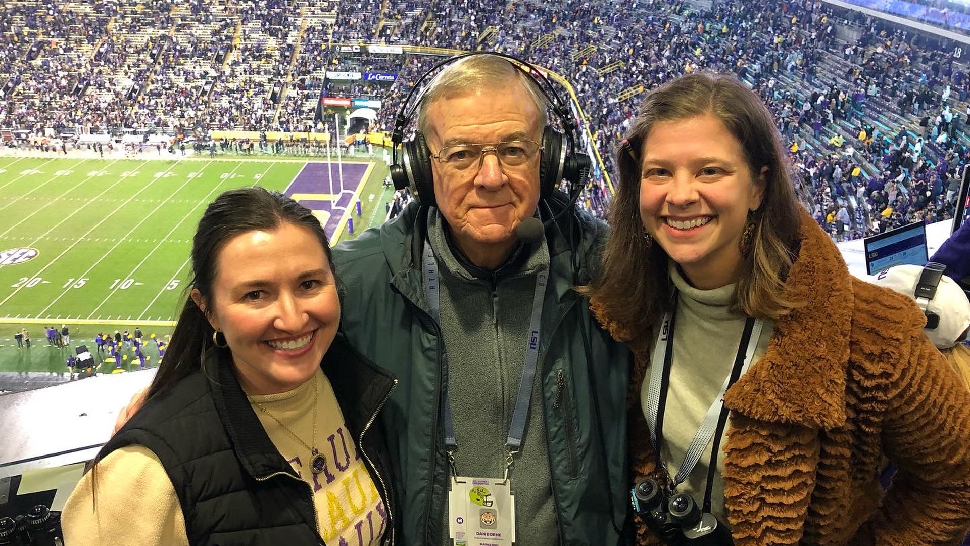 Three people smiling at a crowded football stadium. A man in the center wears a headset and media badge, flanked by two women in jackets, with the field and stands visible behind them.