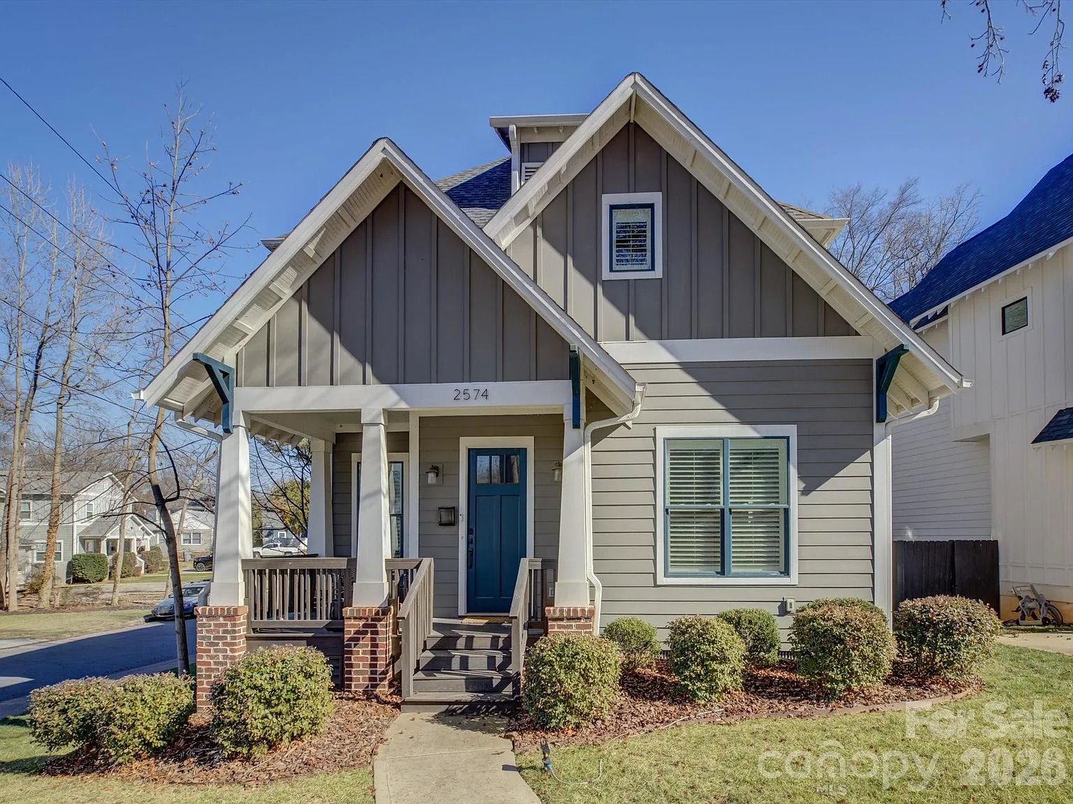 Gray and white two-story house with front porch, blue door, and neatly trimmed bushes under clear blue sky on a sunny day.