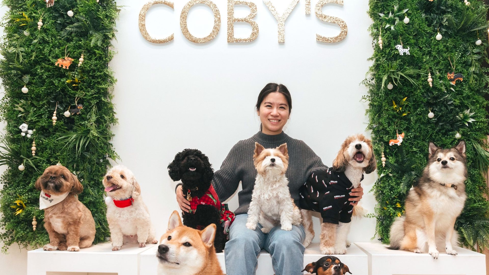 A young woman sits in front of a sign that says "Coby's" while surrounded by dogs.