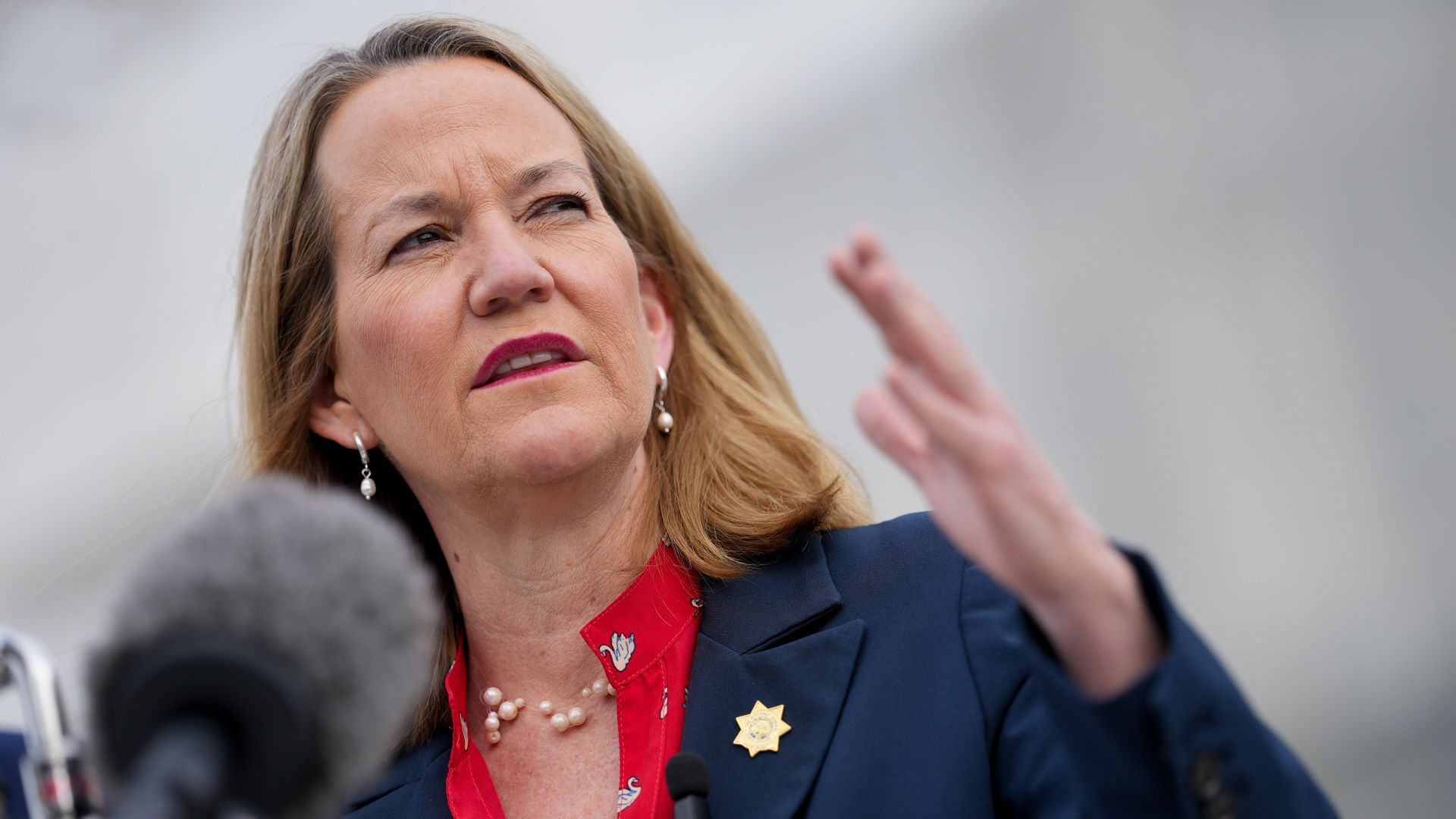 Woman with blonde hair wearing a navy blazer and red shirt with pearl necklace and earrings speaking gesturing with hand, standing in front of microphones against a gray background.