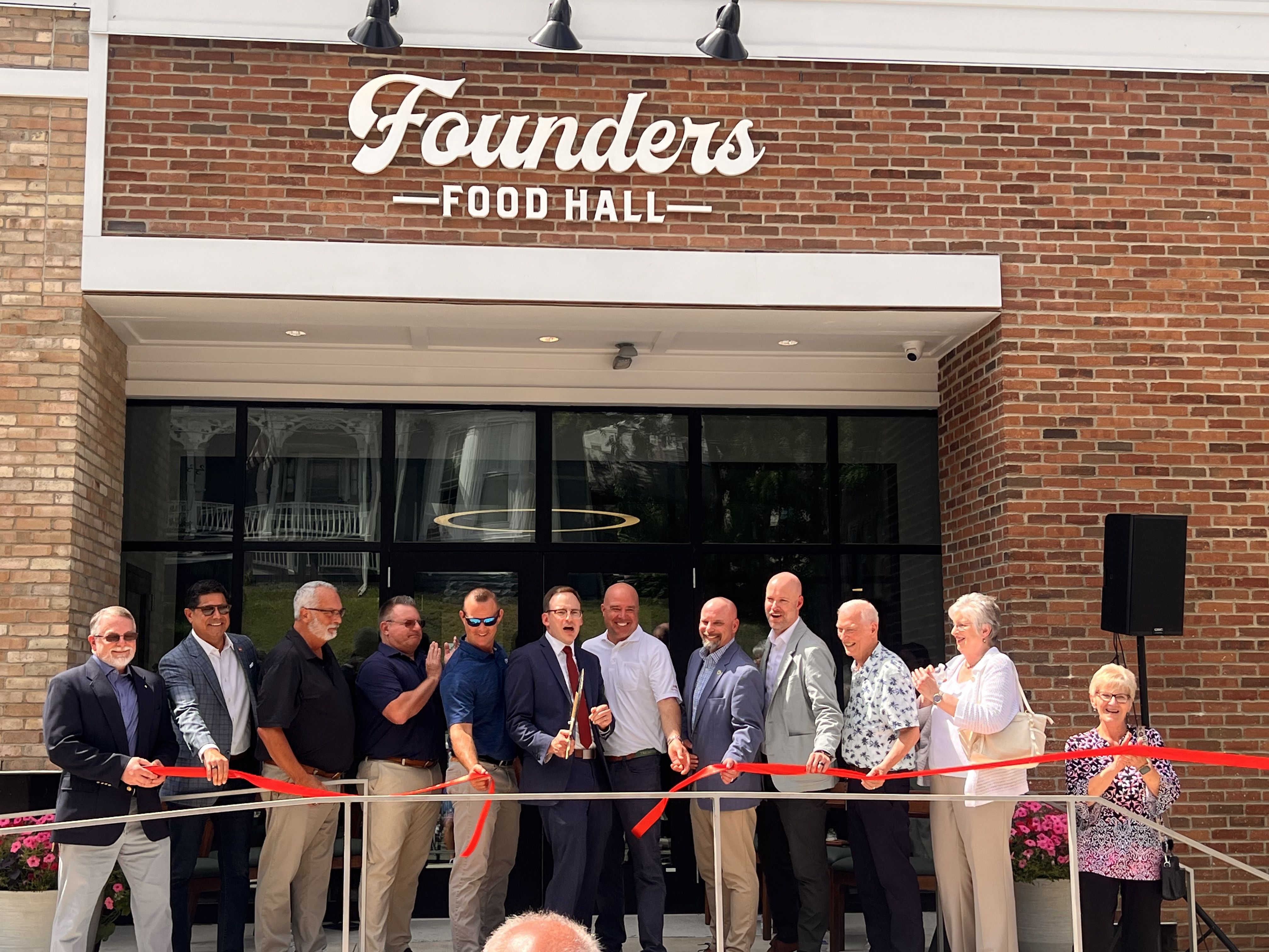 Group of people in business attire cuts a red ribbon in front of a brick building with a sign reading "Founders Food Hall" above glass doors; a ribbon-cutting ceremony in progress.
