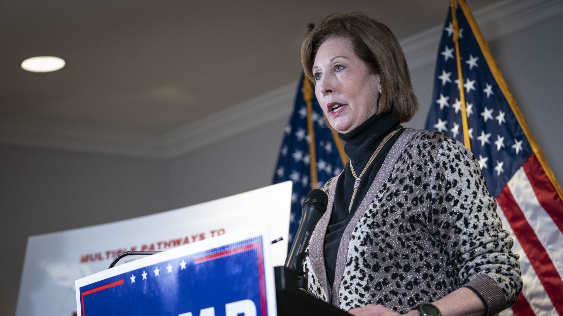 Photo of Sidney Powell speaking from behind a podium with a Trump sign on it