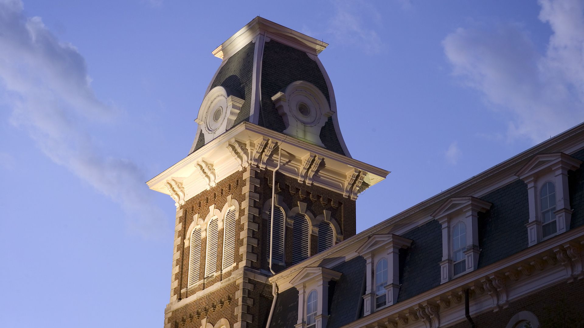 Brick building with a tall tower featuring a curved slate roof, white decorative trim, arched windows, and blue sky with clouds.