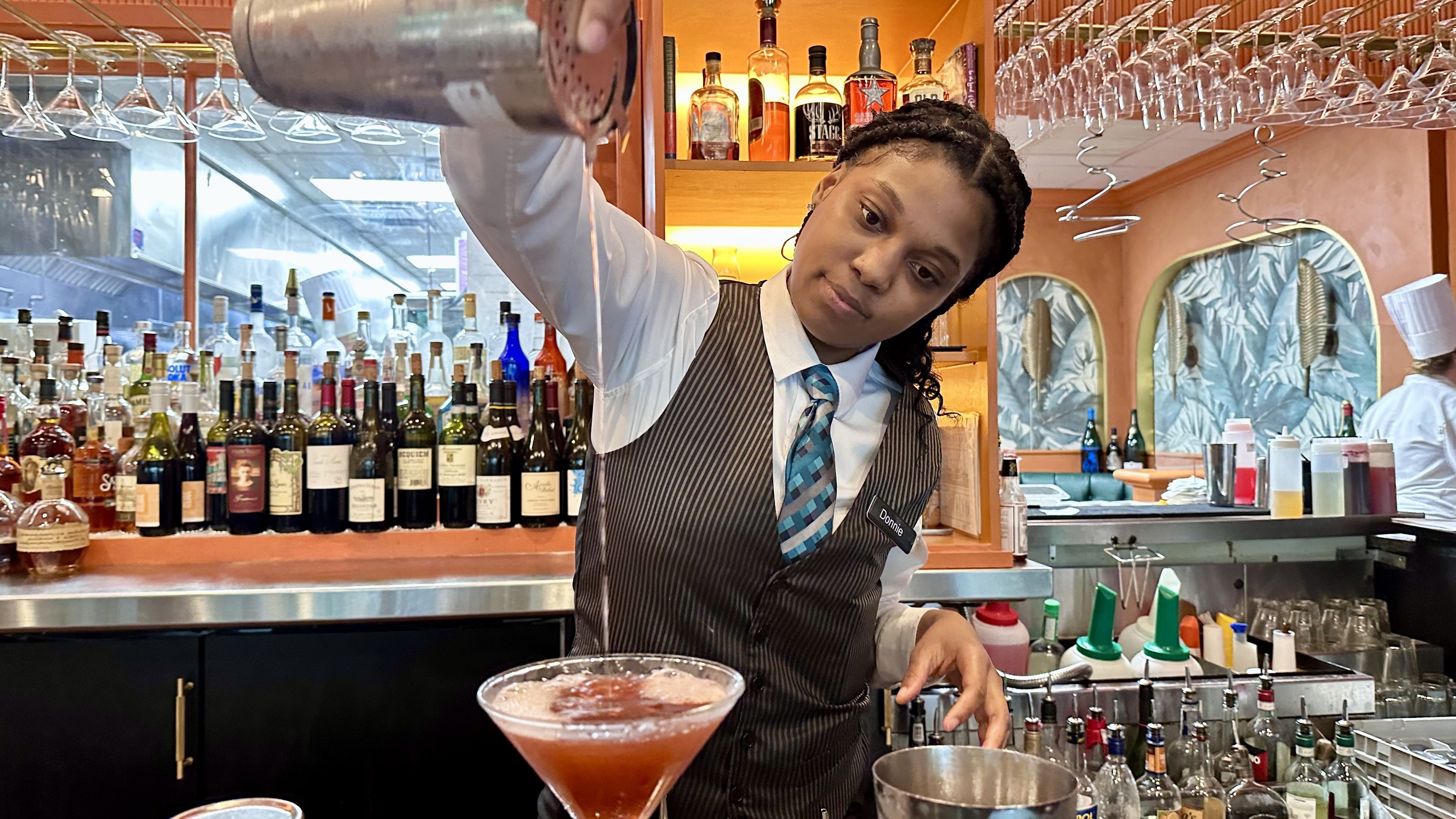 A bartender in a pinstripe vest and teal tie pours a cocktail behind a busy bar with numerous liquor bottles, hanging glasses, and an ice bucket; name tag reads Donnie.