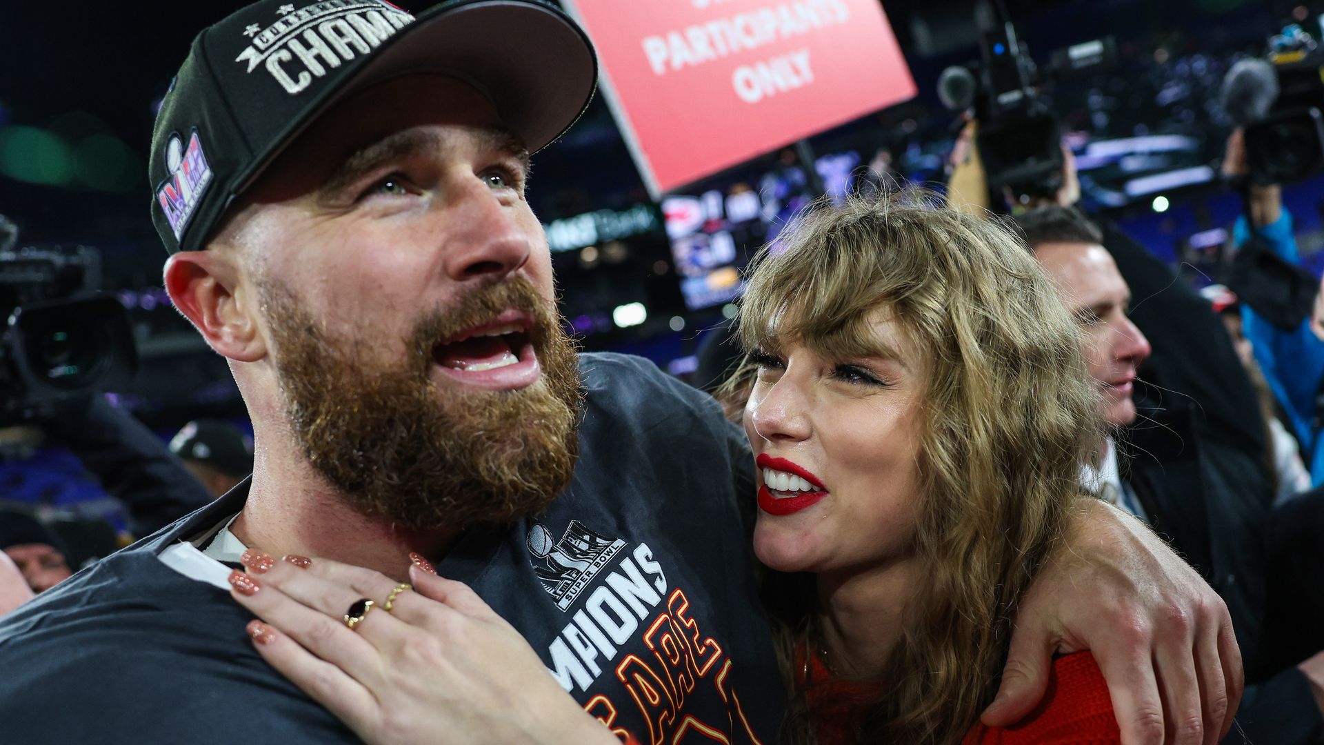 Taylor Swift smiles at and hugs Travis Kelce as they stand on a football field after a game.