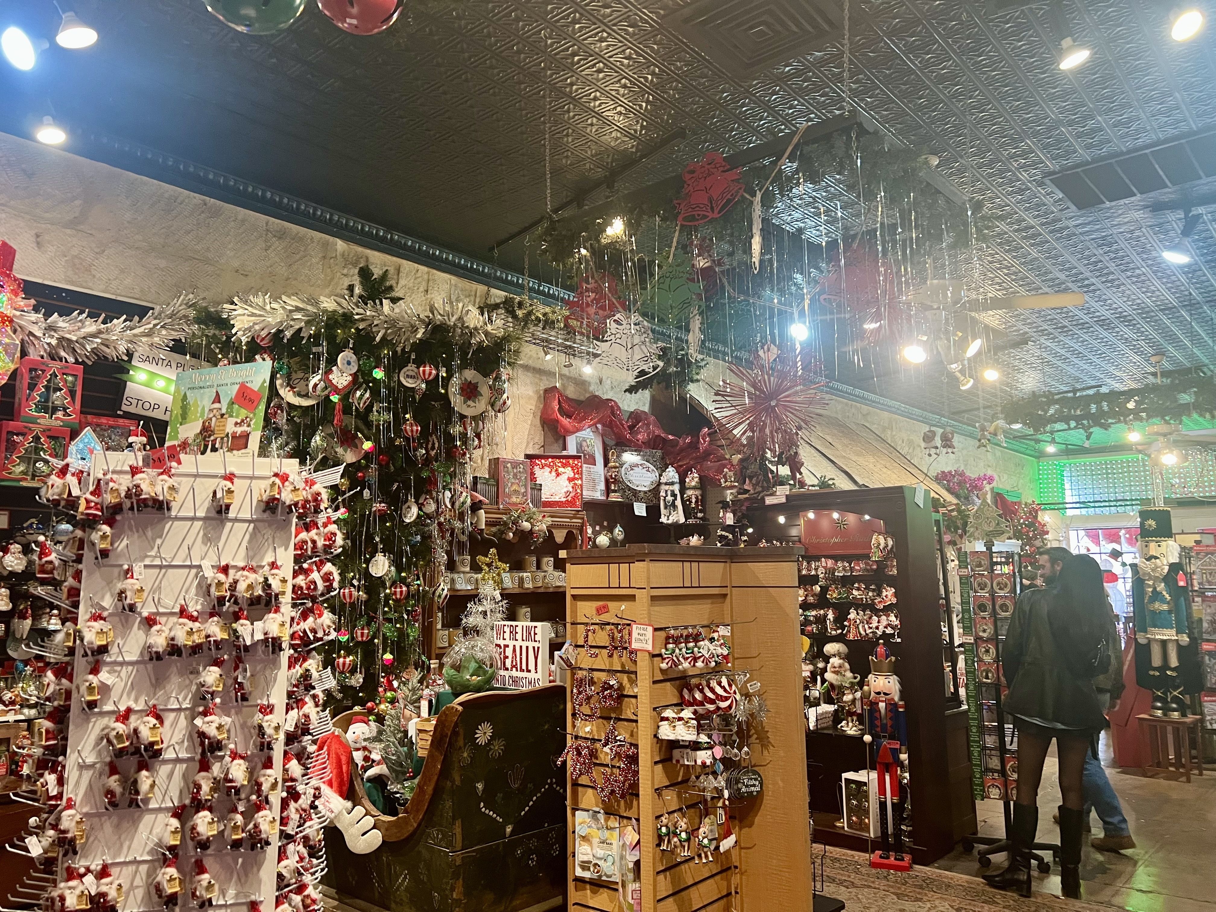 Interior of a festive Christmas shop with decorated trees, ornaments, nutcracker figures, garlands, and hanging tinsel. Two shoppers browse near displays under patterned tin ceiling.