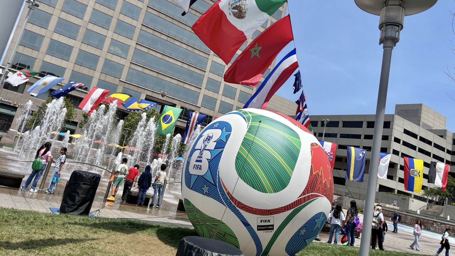 Public plaza with a large colorful FIFA World Cup ball sculpture beside a bubbling fountain; flags from many countries fly above a modern office building as people walk by.