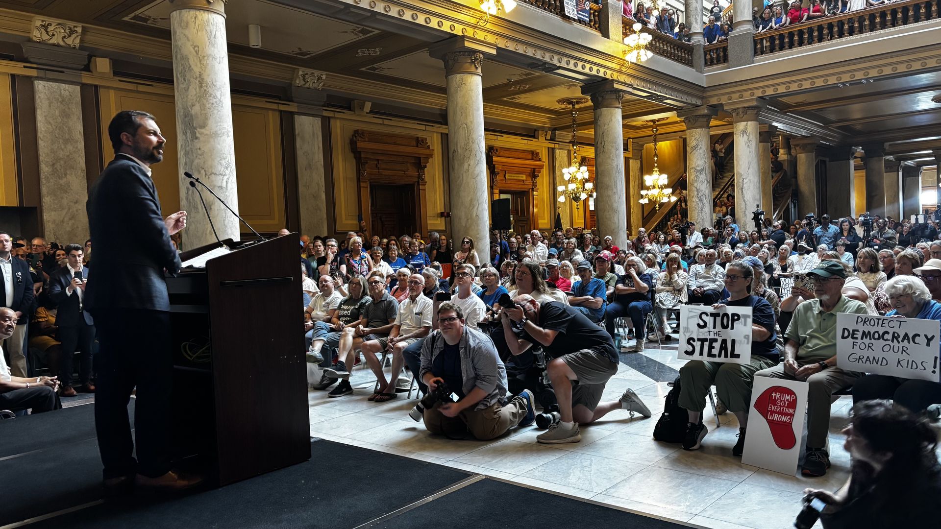 Man in dark suit speaking at podium in ornate hall with marble columns, large crowd seated and standing, some holding signs reading "STOP THE STEAL" and "PROTECT DEMOCRACY FOR OUR GRAND KIDS".