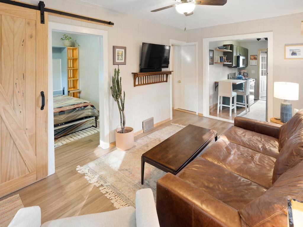 A living room with white walls, a leather couch, a tv on the wall and an open barn door showing a bedroom.