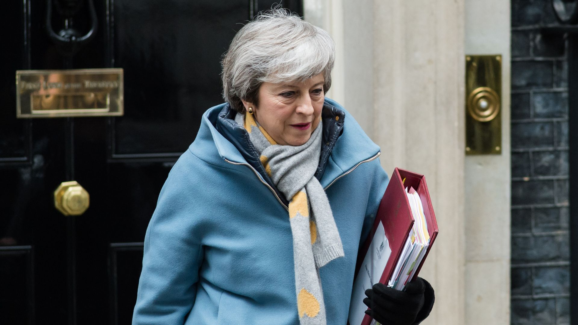 Theresa May walks out of a black doorway while holding a binder and wearing a winter coat.
