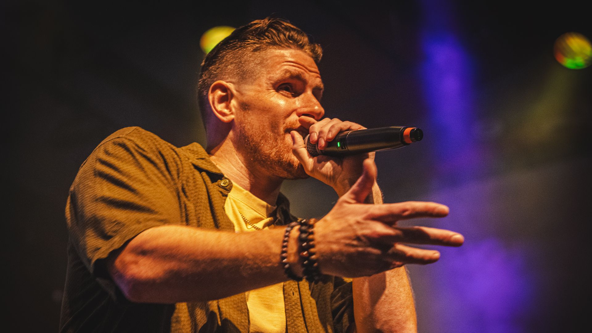 A picture of Cane onstage with holding a mic close to his mouth with his right arm outstretched.
