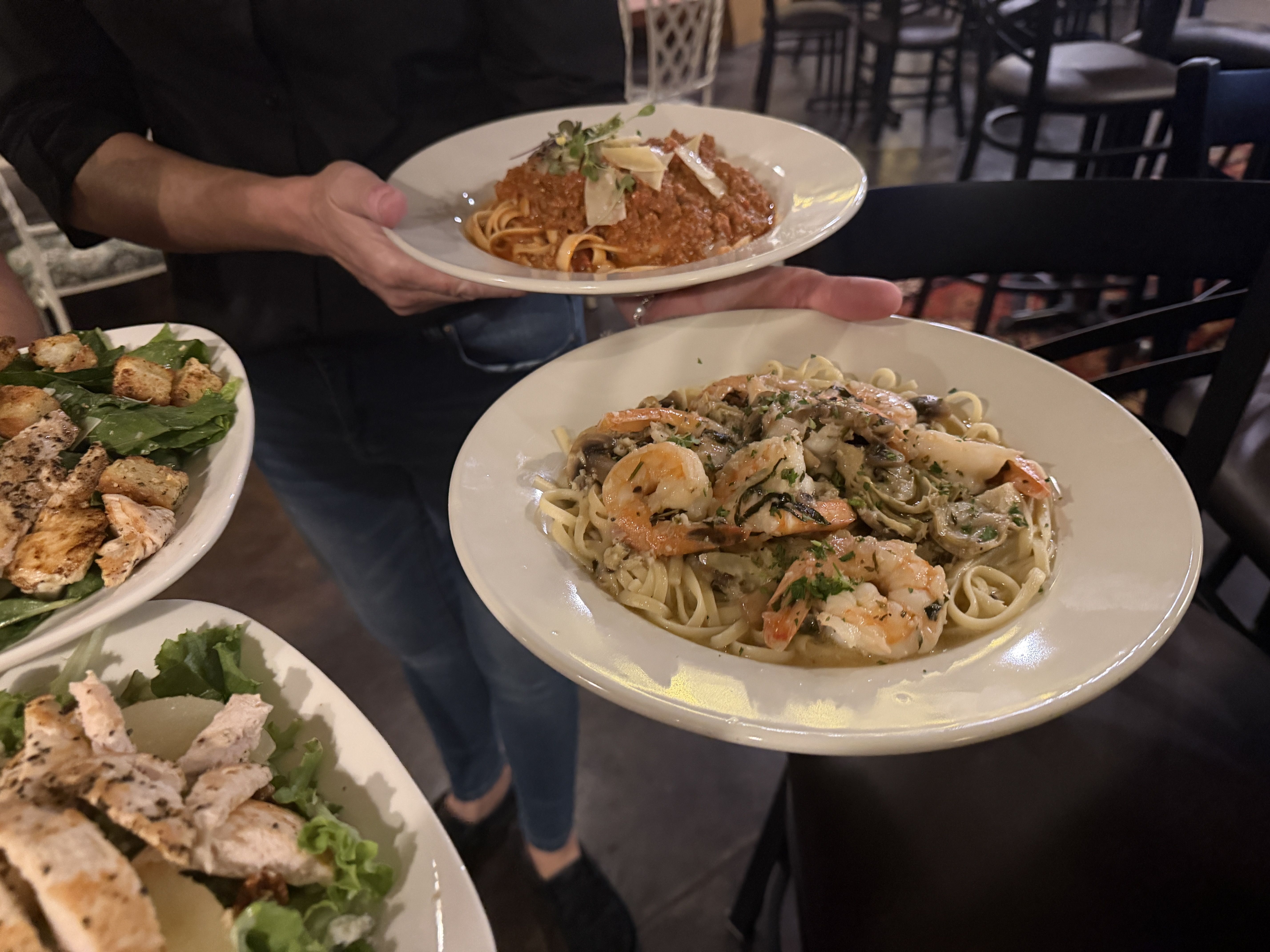 Server in black holds a plate of spaghetti with meat sauce, shaved parmesan, and microgreens; in the foreground, a shrimp-mushroom pasta in cream sauce and a greens salad with croutons.