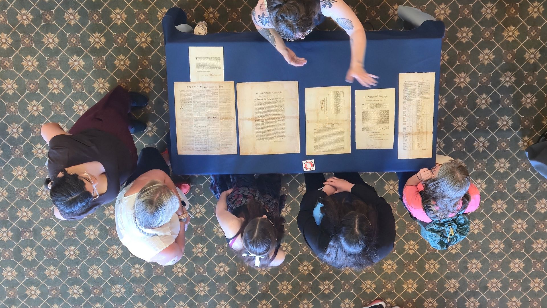Overhead view of a blue table with several old, yellowed documents laid out in a row; five people lean over the table on a patterned carpet, examining the papers.