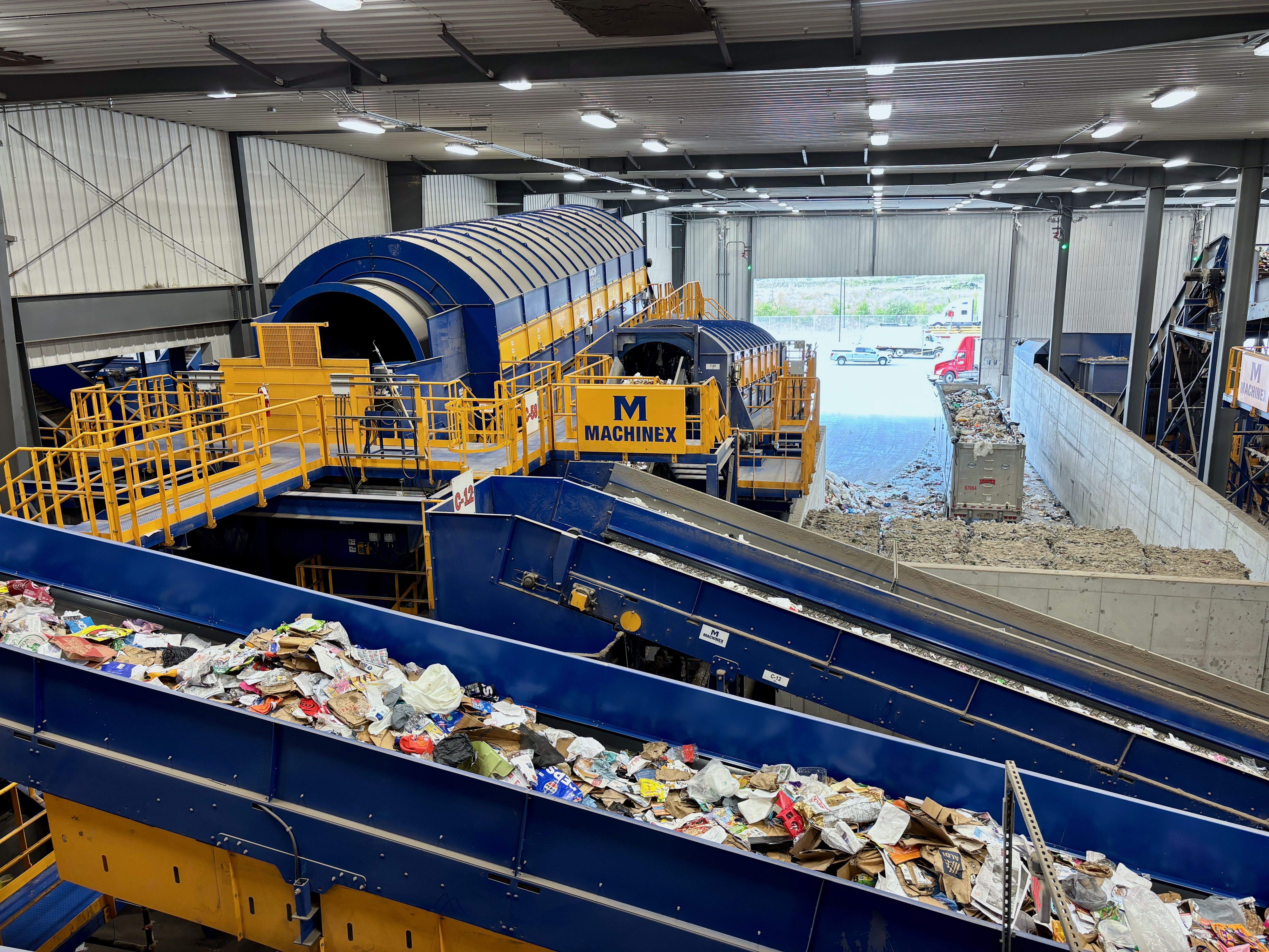 Inside a waste recycling plant, large blue and yellow machinery with a rotating cylindrical duct and yellow safety rails; conveyor belts carry piles of mixed trash under bright overhead lights.